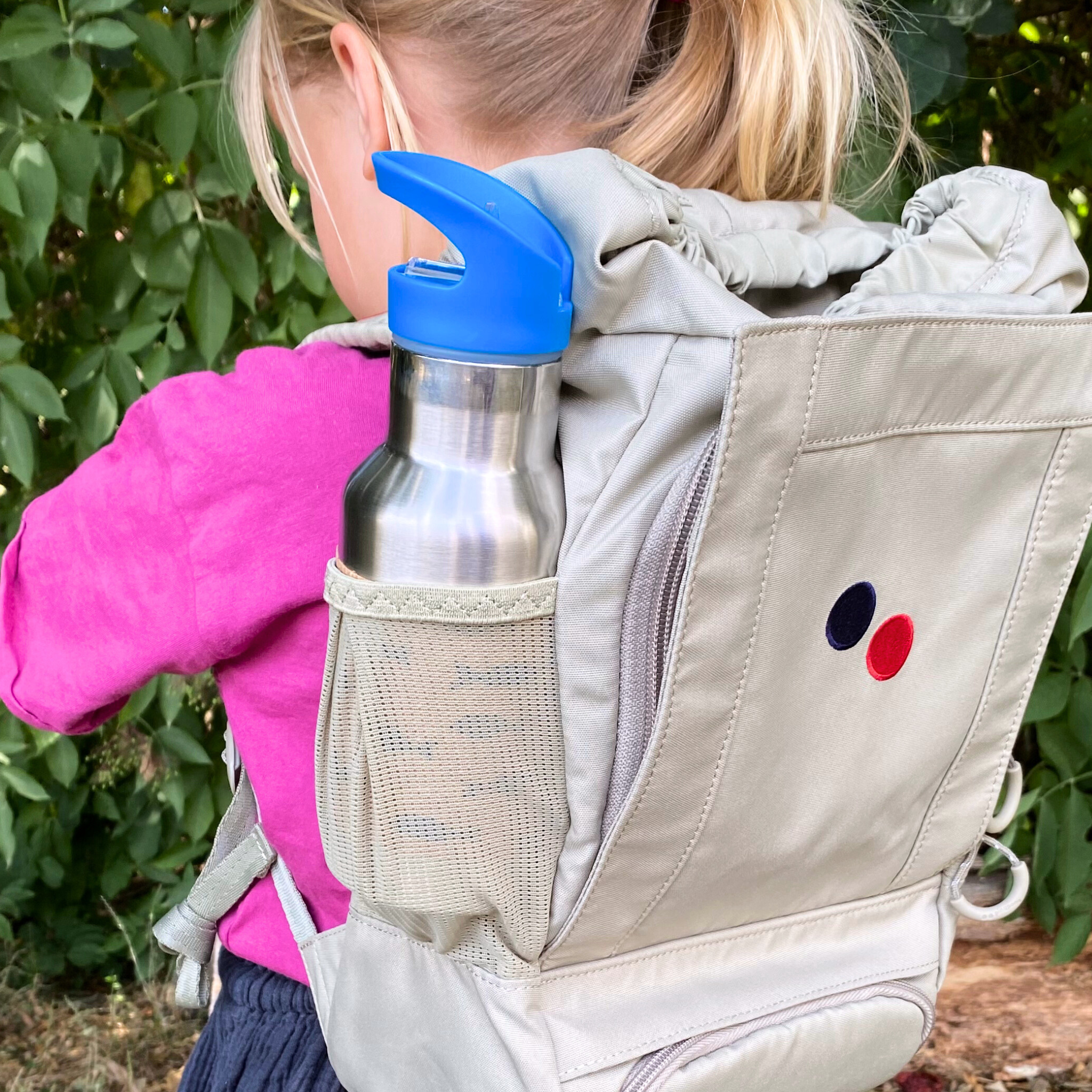 Ein kleines Kind mit blondem Haar mit einem Pferdeschwanz trägt einen hellbeigen Rucksack mit einer blauen Wasserflasche in der Seitentasche. Das Kind steht im Freien vor einem grünen Gebüsch.