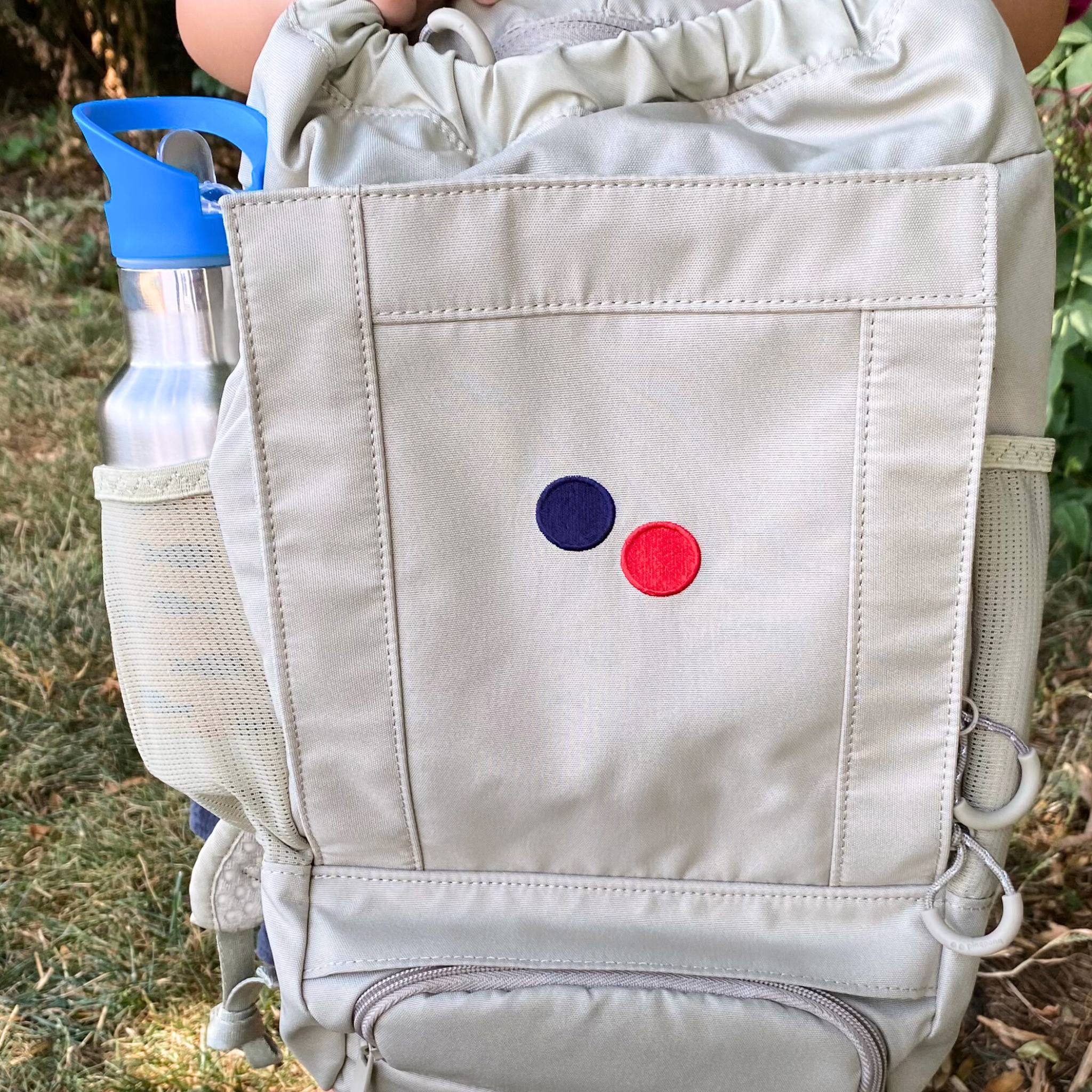 Ein hellgrauer Rucksack mit einem blauen und einem roten Punkt, die auf die Vordertasche gestickt sind. In der seitlichen Netztasche befindet sich eine Wasserflasche aus Metall mit blauem Deckel, und der Rucksack steht im Freien auf Gras.