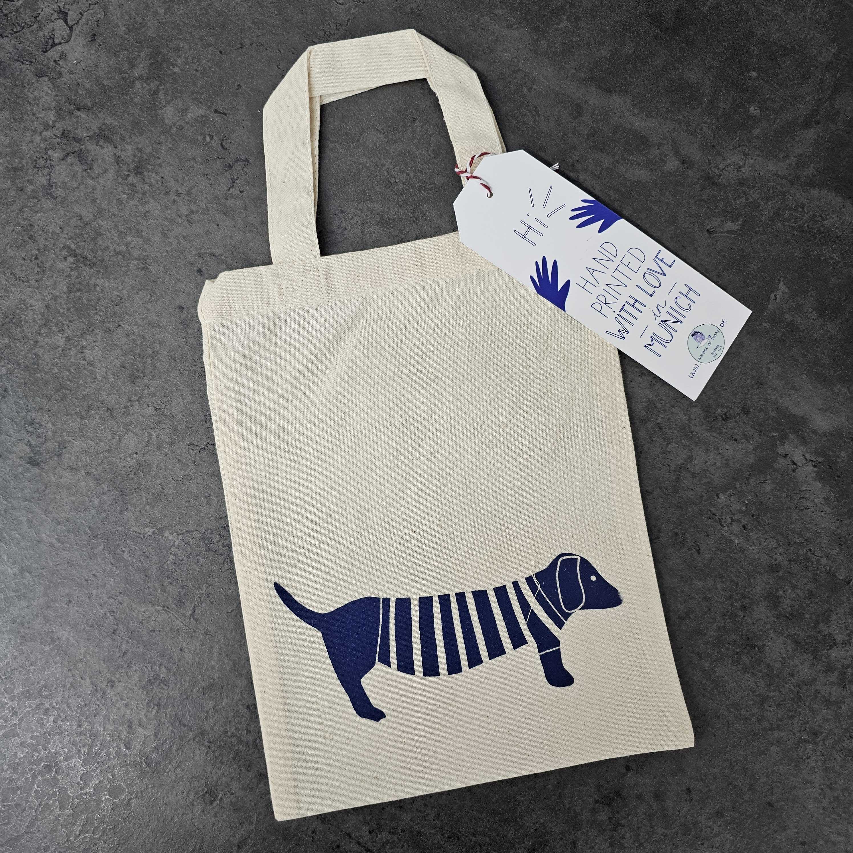 Eine cremefarbene Tragetasche mit einem blauen Aufdruck eines Dackels, der einen gestreiften Pullover trägt. Auf einem am Henkel befestigten Schildchen steht: Hi! Handbedruckt mit Liebe in Notts, Munch. Die Tasche ist auf einem dunklen Stein Oberfläche.