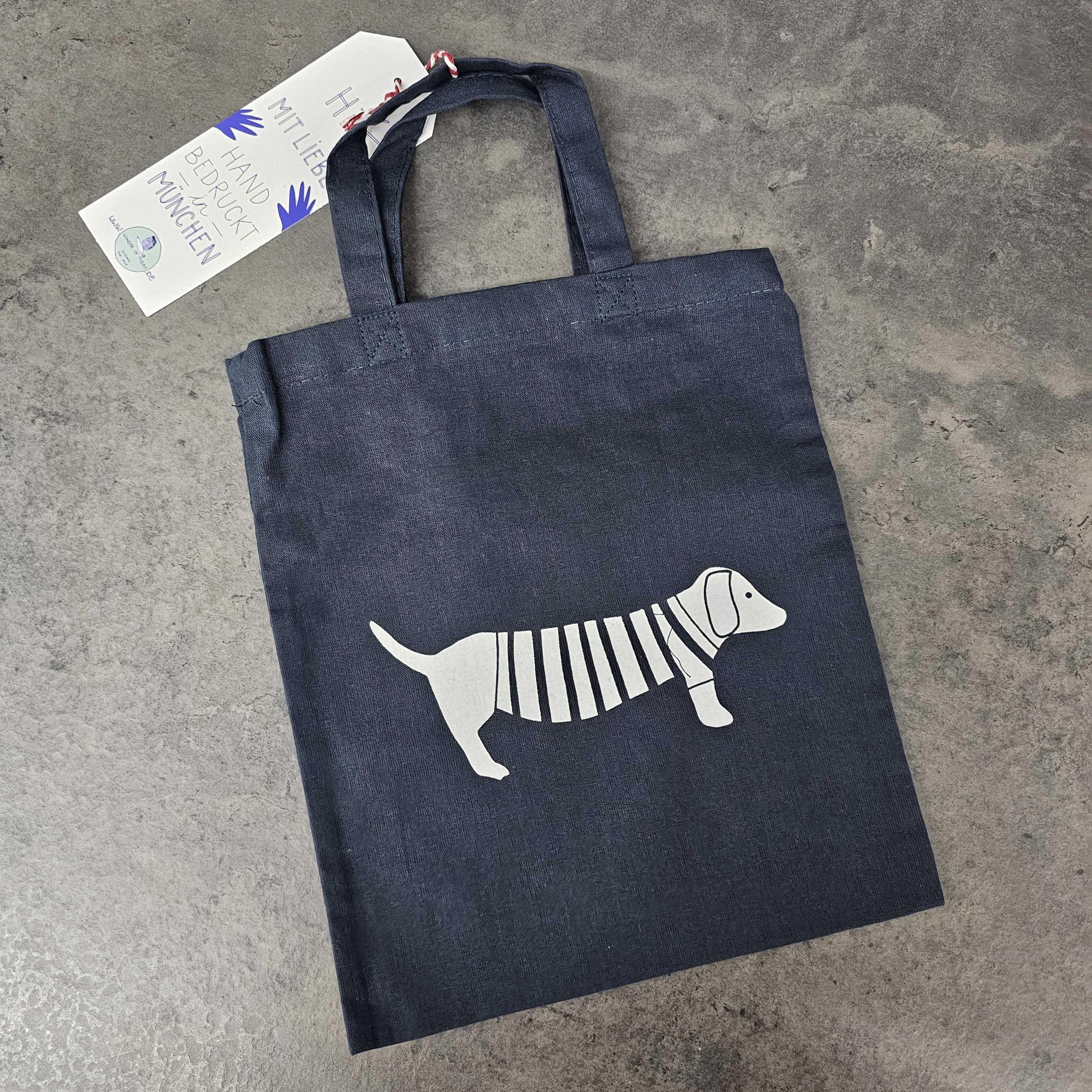 Eine dunkelblaue Tragetasche mit Henkeln, auf der eine weiße Illustration eines Dackels mit gestreiftem Pullover abgebildet ist. Die Tasche steht auf einer grauen Betonfläche, und an einem Henkel ist ein Anhänger befestigt.