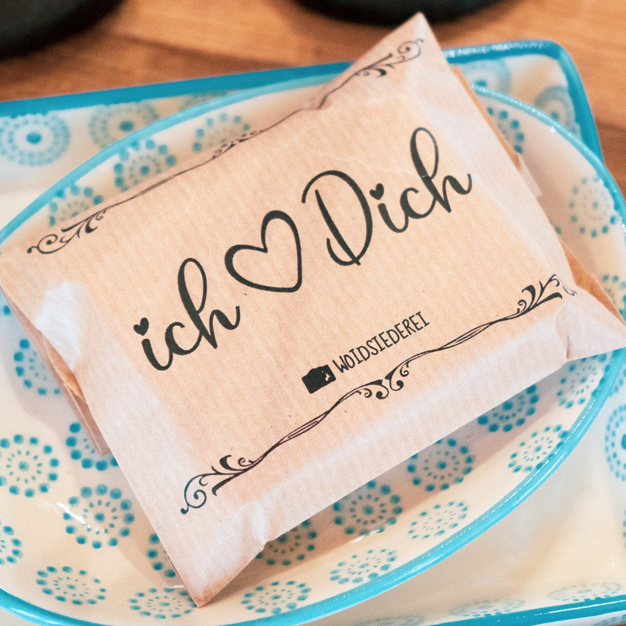 Ein kleines braunes Papierpaket mit dem Aufdruck "Ich ♥ Dich", das in einer dekorativen blau-weißen Schale auf einer Holzfläche liegt.