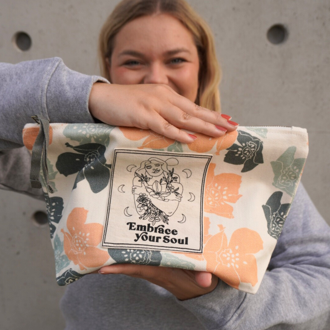 Frau hält eine Tasche mit floralem Muster und der Aufschrift "Embrace Your Soul" vor einem strukturierten Hintergrund.
