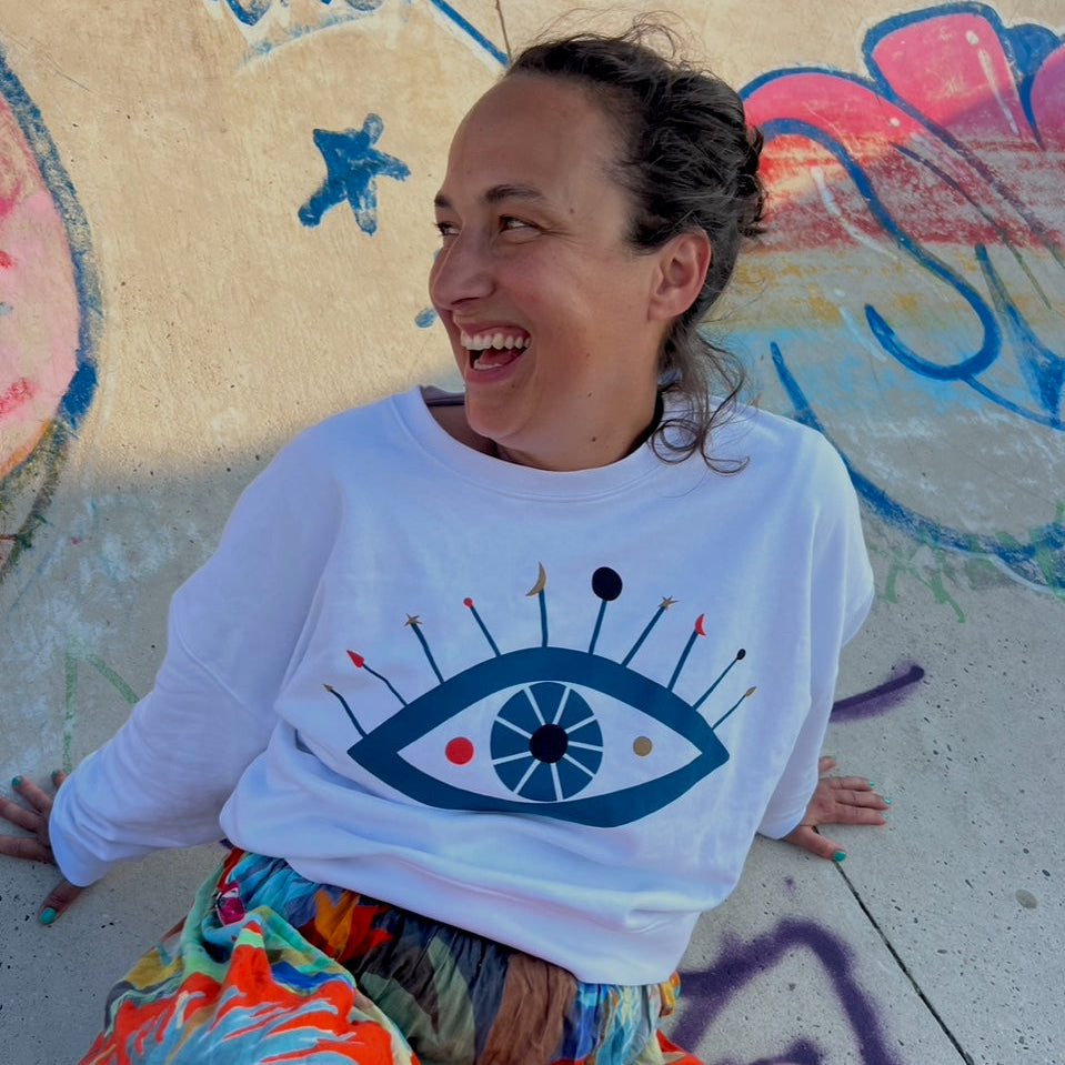 Eine lächelnde Person mit lockigem Haar sitzt auf dem Boden und lehnt sich zurück. Sie trägt ein weißes Sweatshirt mit einer großen Augengrafik und einen bunten Rock. Der Hintergrund besteht aus lebhaften Graffiti und gemalten Formen auf Beton.