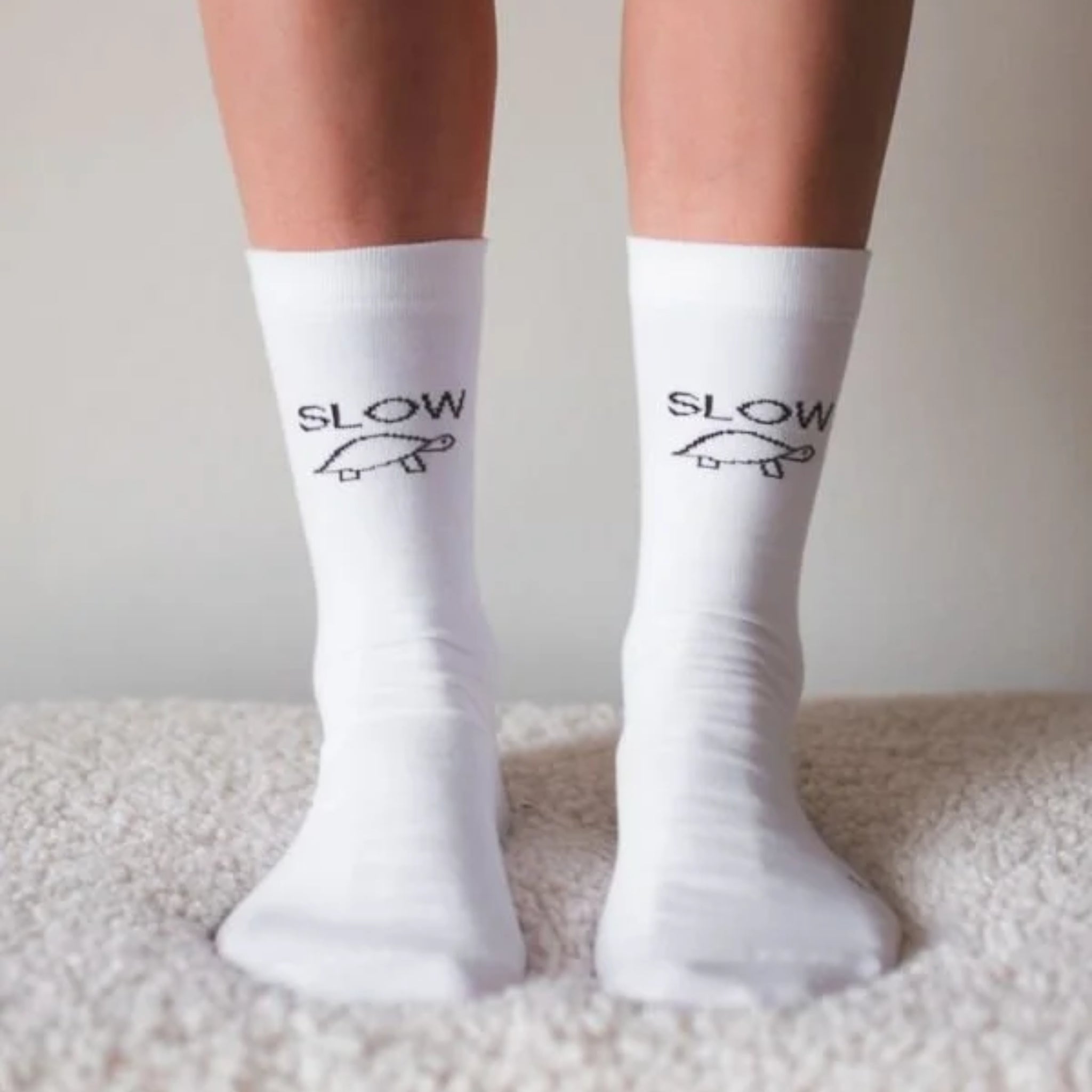 Eine Person, die weiße Socken mit dem Wort SLOW und einer einfachen Schildkrötenillustration auf jeder Socke trägt und auf einer weichen, strukturierten Oberfläche steht.