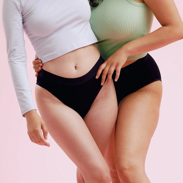 Zwei eng beieinander stehende Frauen vor einem hellrosa Hintergrund, die hochtaillierte schwarze Unterwäsche und pastellfarbene Oberteile tragen. Nur ihre Körper sind von den Schultern bis zu den Oberschenkeln zu sehen.