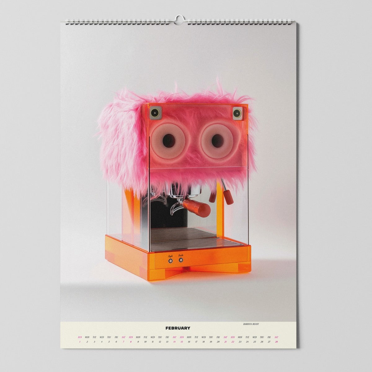 Ein Kalenderblatt für Februar zeigt eine skurrile Kaffeemaschine, die mit leuchtend pinkem Kunstfell und großen Kulleraugen verziert ist, was ihr ein verspieltes, monstermäßiges Aussehen verleiht.
