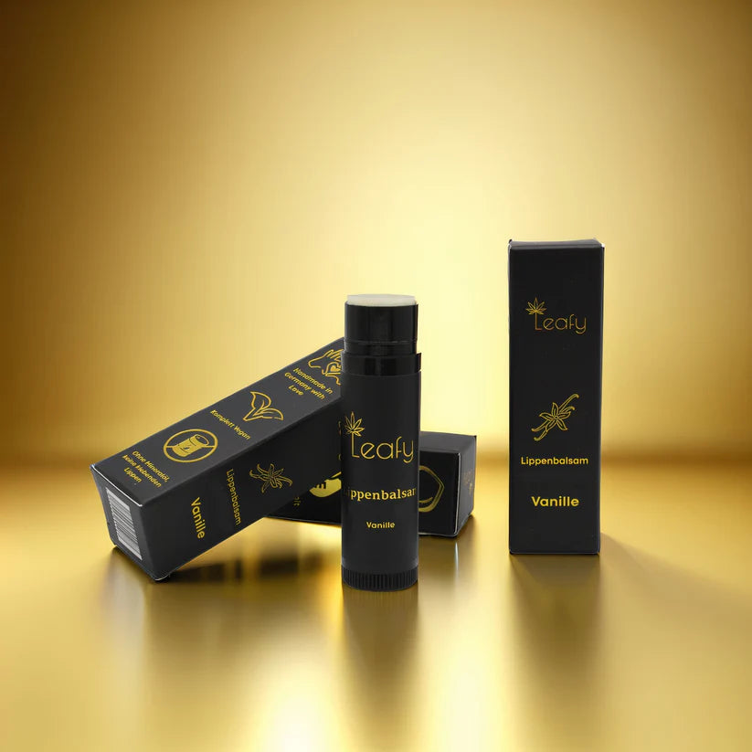 Eine schwarze Tube Leafy Vanille-Lippenbalsam steht aufrecht neben der dazugehörigen Schachtel, zwei weitere Schachteln liegen schräg dahinter, alles vor einem glänzenden Goldhintergrund.