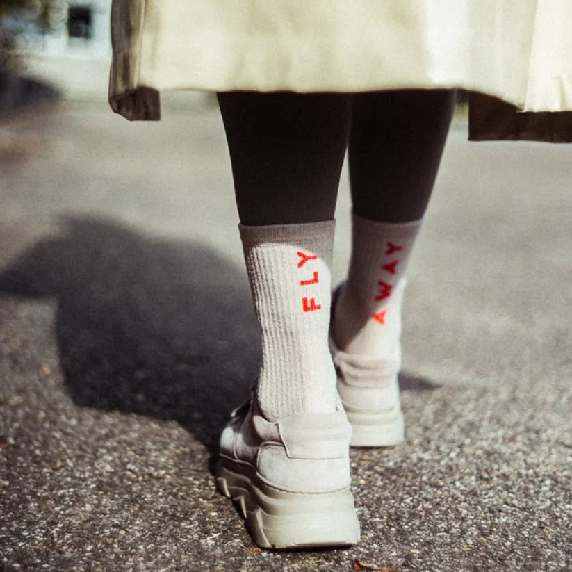 Person, die einen beigen Mantel, schwarze Leggings, weiße Plateauschuhe und Socken mit der Aufschrift "FLY AWAY" in roten Buchstaben trägt und auf einem sonnenbeschienenen Bürgersteig steht. Das Bild ist von hinten in Bodennähe aufgenommen.
