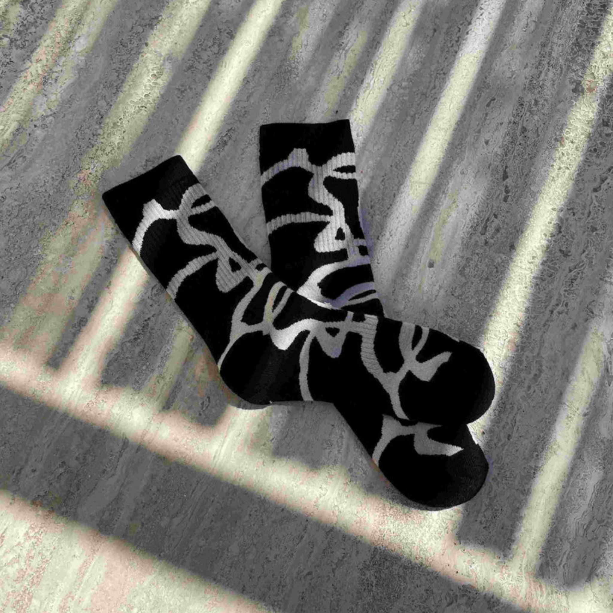 Ein Paar schwarze Socken mit kühnen weißen abstrakten Mustern liegt auf einer strukturierten grauen Oberfläche, die von gestreiftem Sonnenlicht und Schatten beleuchtet wird.