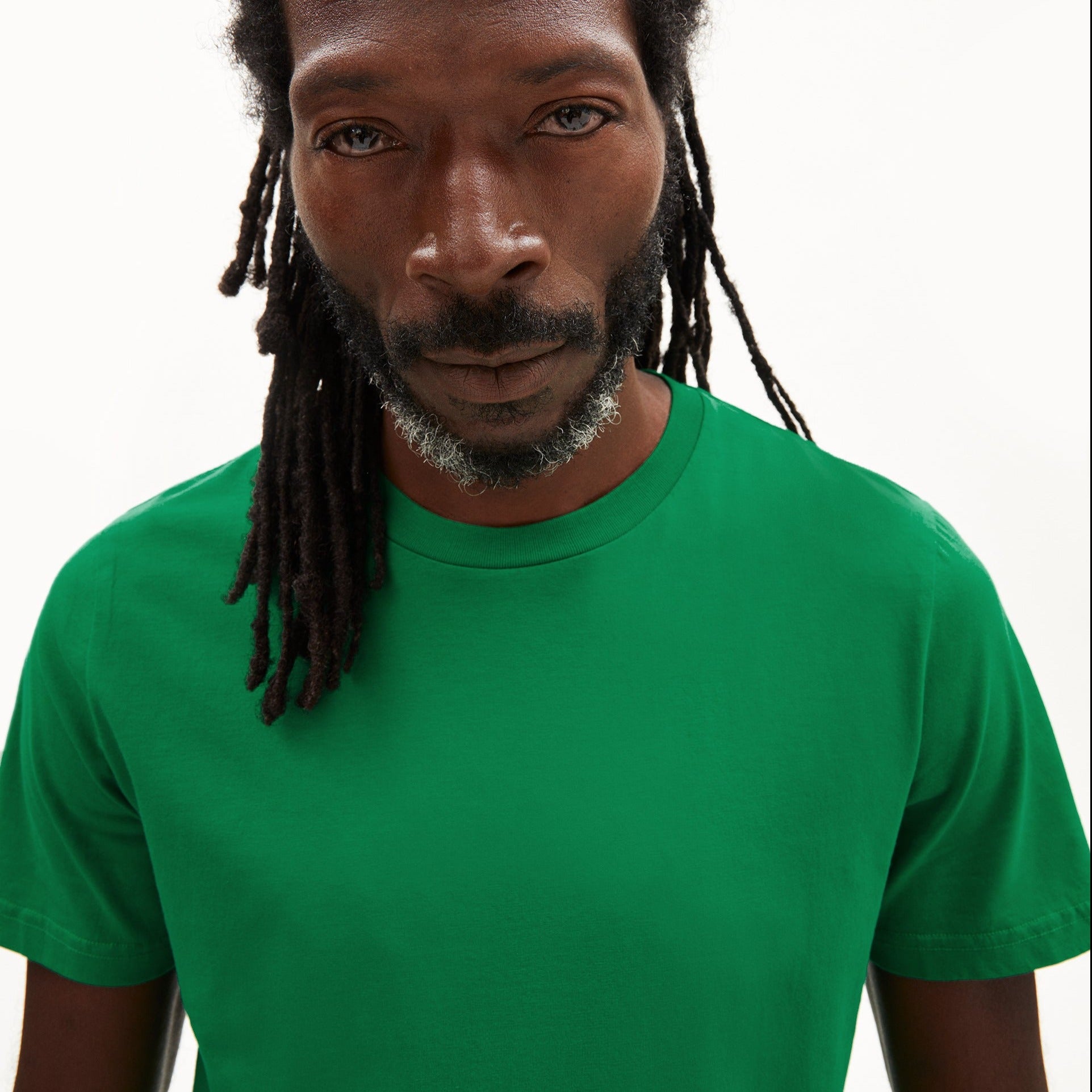 Mann mit langen Dreadlocks trägt ein grünes T-Shirt und blickt ernst in die Kamera vor einem weißen Hintergrund.