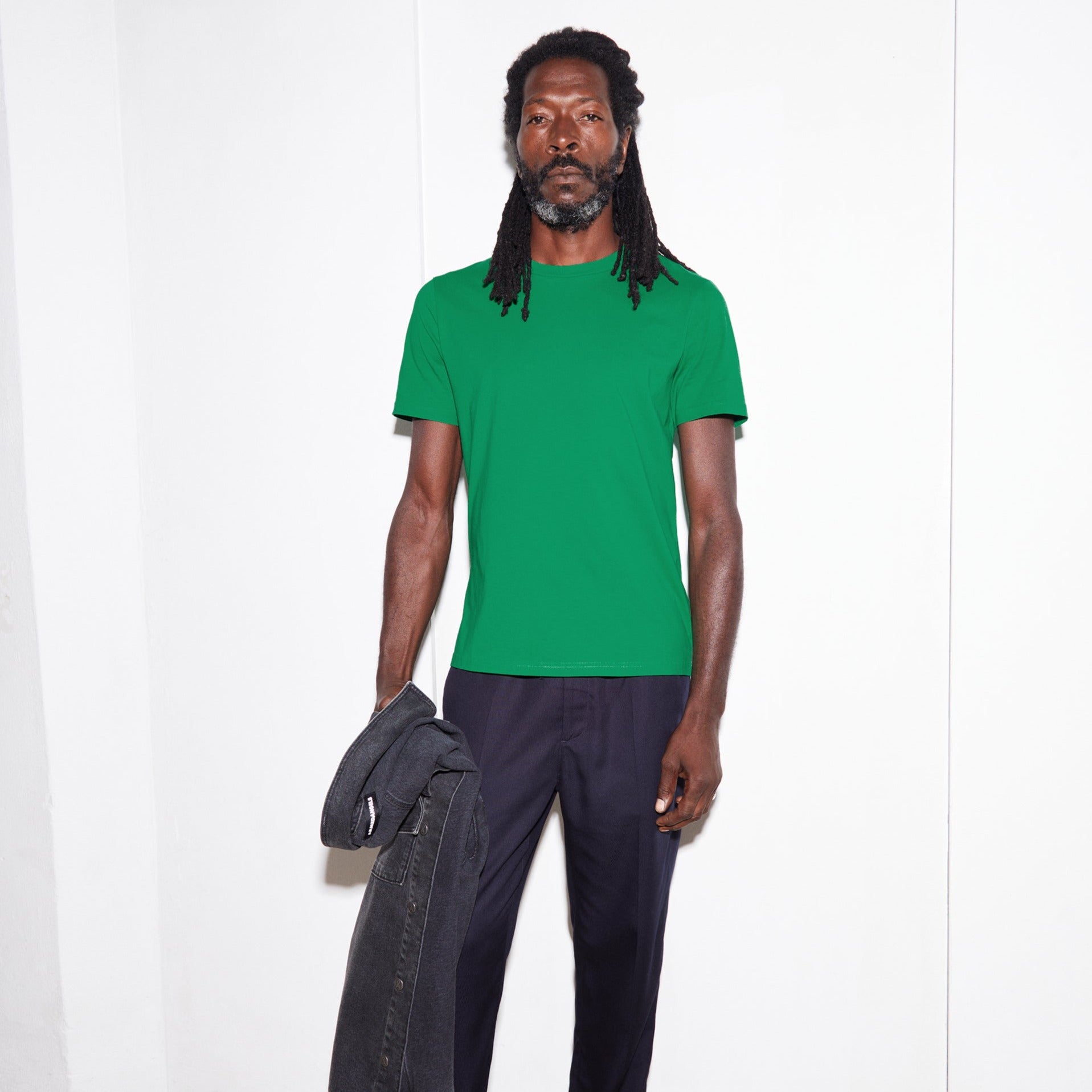 Person mit Dreadlocks trägt ein grünes T-Shirt und schwarze Hose, hält eine schwarze Jacke und steht vor weißer Wand.