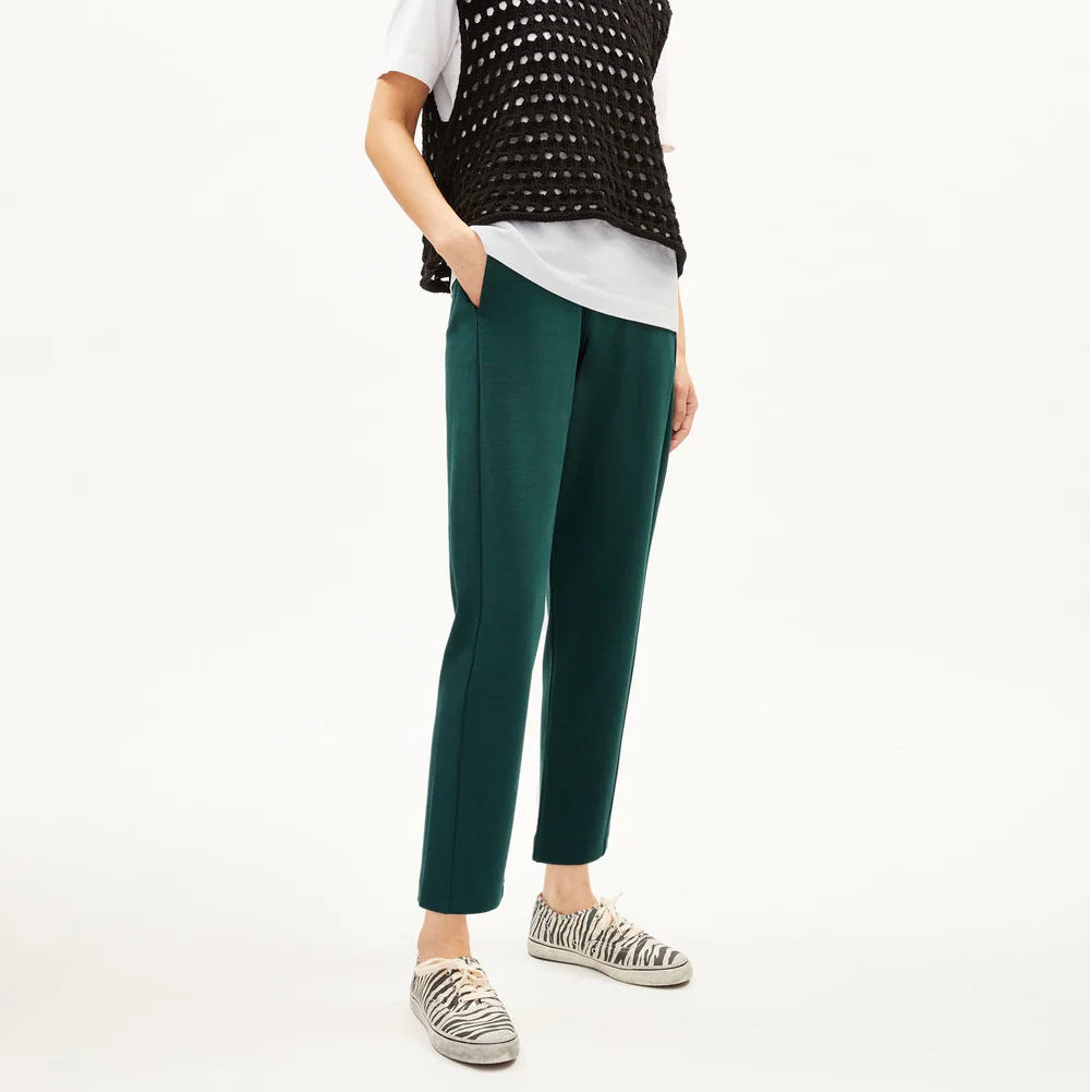 Eine Person steht vor einem schlichten Hintergrund und trägt eine schwarze Netzweste über einem weißen T-Shirt, eine blaugrüne Hose, Slipper mit Zebradruck und eine schwarze Baseballkappe. Ihre Hände sind in den Taschen und sie schaut nach vorne.