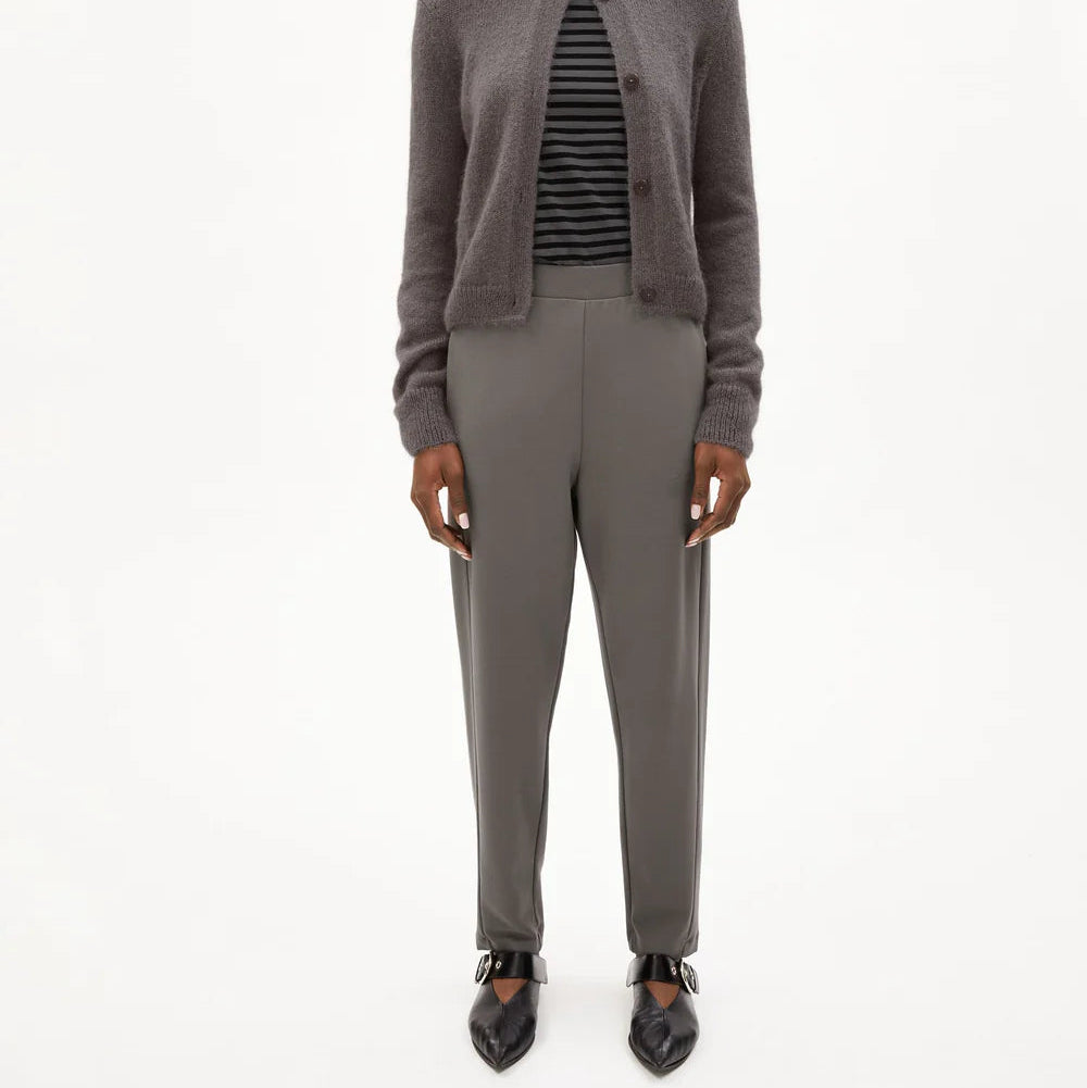 Eine Frau steht vor einem schlichten weißen Hintergrund und trägt eine graue Mütze, eine graue Strickjacke mit Knöpfen über einem schwarz-grau gestreiften Rollkragenpullover, eine graue Hose mit hoher Taille und schwarze spitze Schuhe.