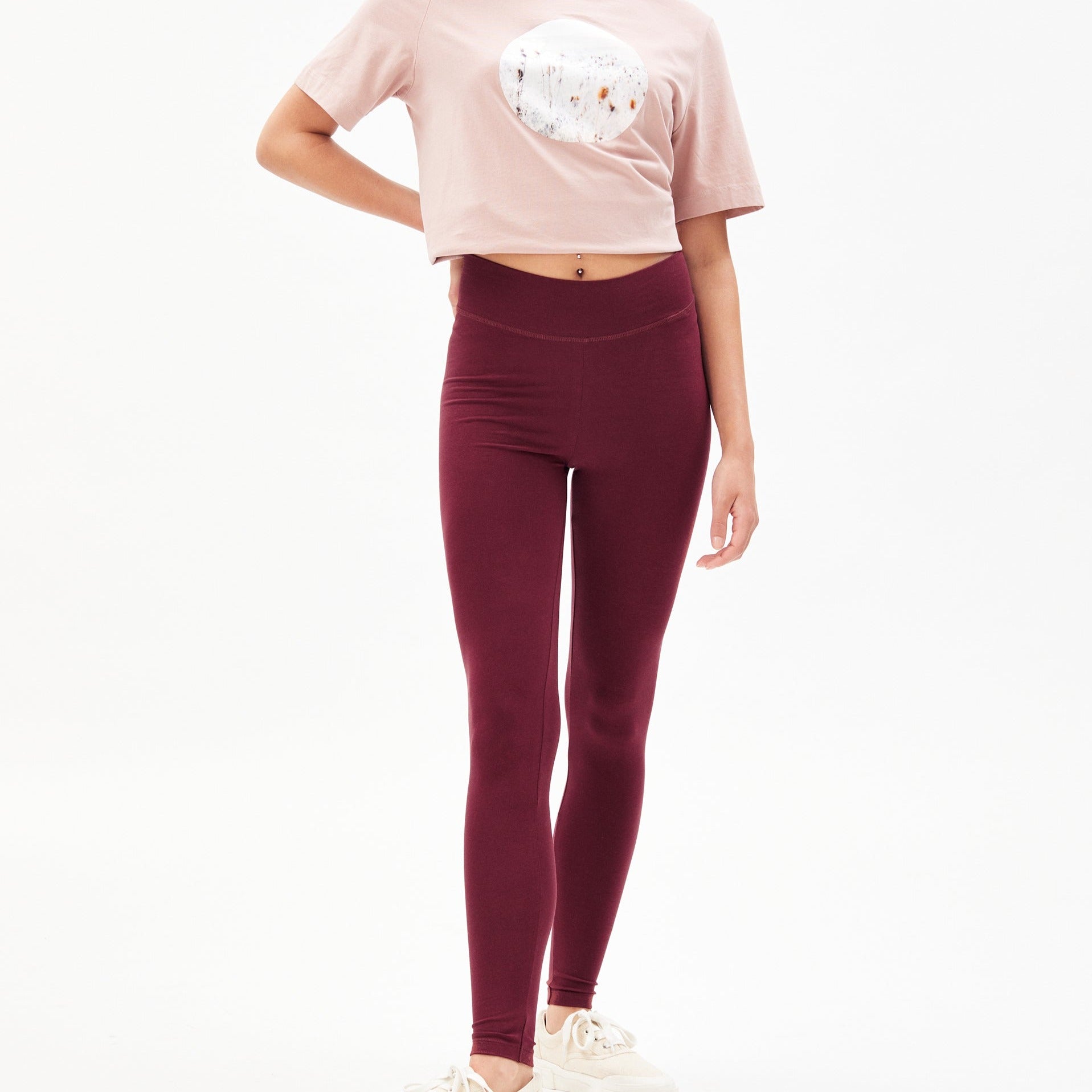 Person trägt ein rosafarbenes T-Shirt und weinrote Leggings, eine Hand auf der Hüfte, vor weißem Hintergrund.