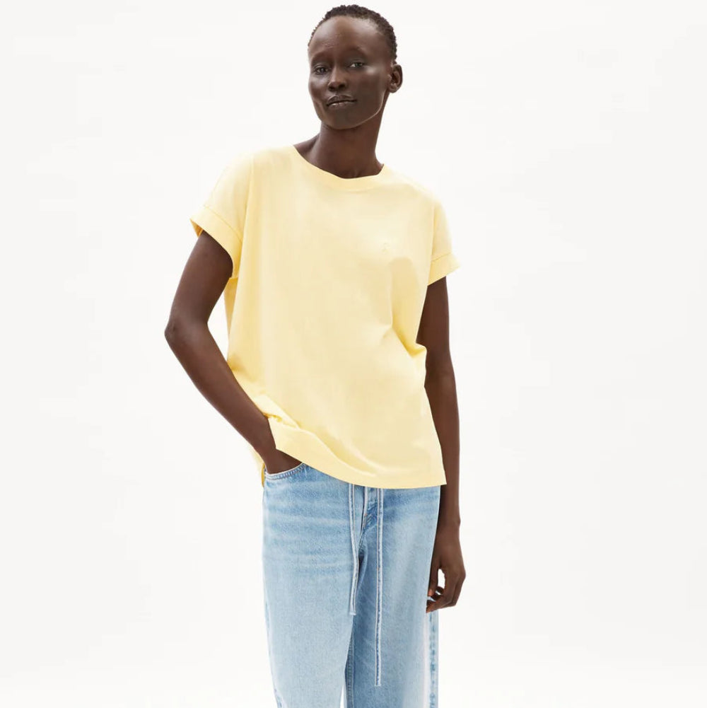 Eine Person steht vor einem schlichten weißen Hintergrund, trägt ein lockeres gelbes T-Shirt und hellblaue, weit geschnittene Jeans mit weißen Schuhen. Eine Hand steckt in ihrer Tasche und sie lächelt sanft.