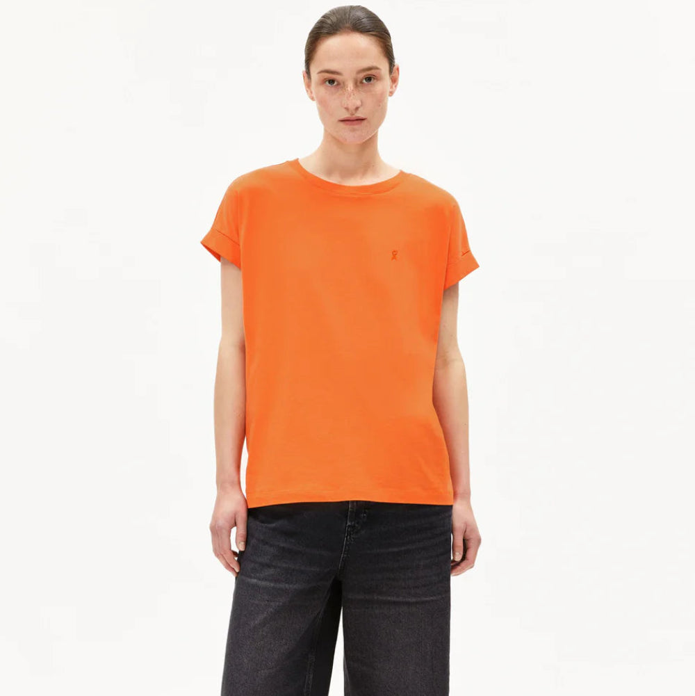 Eine Person steht vor einem schlichten weißen Hintergrund und trägt ein orangefarbenes T-Shirt, knielange schwarze Jeans-Shorts und schwarze Sandalen mit roten Riemen.