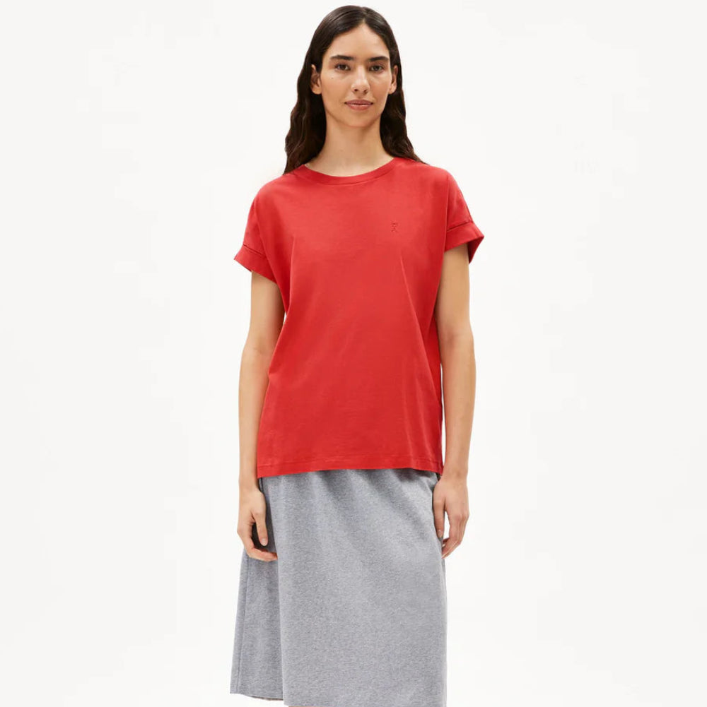 Eine Frau steht mit dem Gesicht nach vorne, trägt ein lockeres rotes T-Shirt, einen knielangen grauen Rock und schwarze Slide-Sandalen vor einem schlichten weißen Hintergrund.