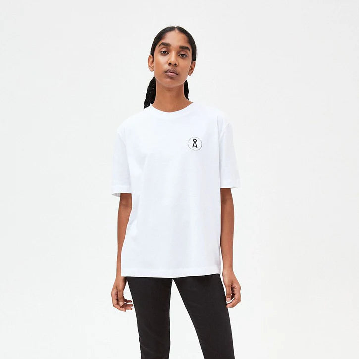 Eine Person steht vor einem schlichten weißen Hintergrund und trägt ein weißes T-Shirt mit einem kleinen runden Logo auf der linken Brust, eine schwarze Hose und weiße Turnschuhe. Sie hat lange dunkle Haare, die nach hinten gezogen sind.