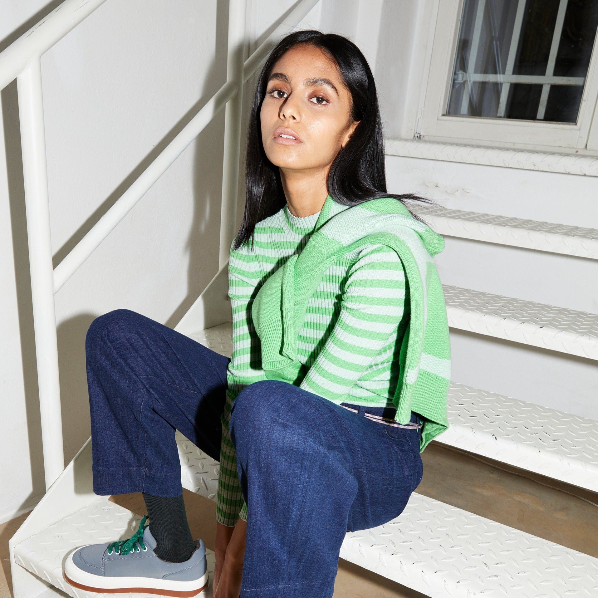 Frau in gestreiftem grünen Pullover und blauer Hose sitzt auf einer Treppe, mit einem grünen Pullover über den Schultern.