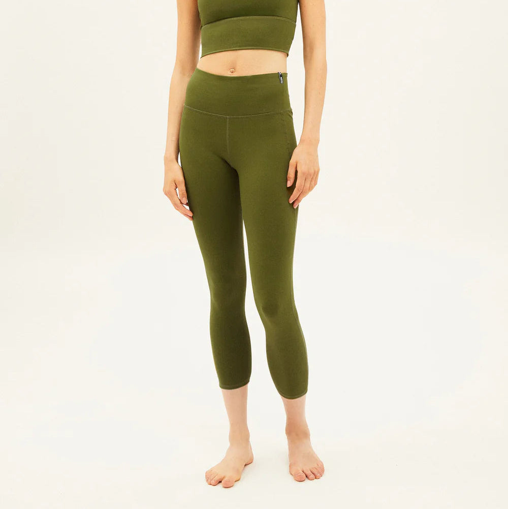 Eine Frau steht barfuß auf einem schlichten Hintergrund und trägt einen olivgrünen Sport-BH und dazu passende hoch taillierte Leggings. Sie blickt mit neutralem Blick und entspannter Körperhaltung direkt in die Kamera.