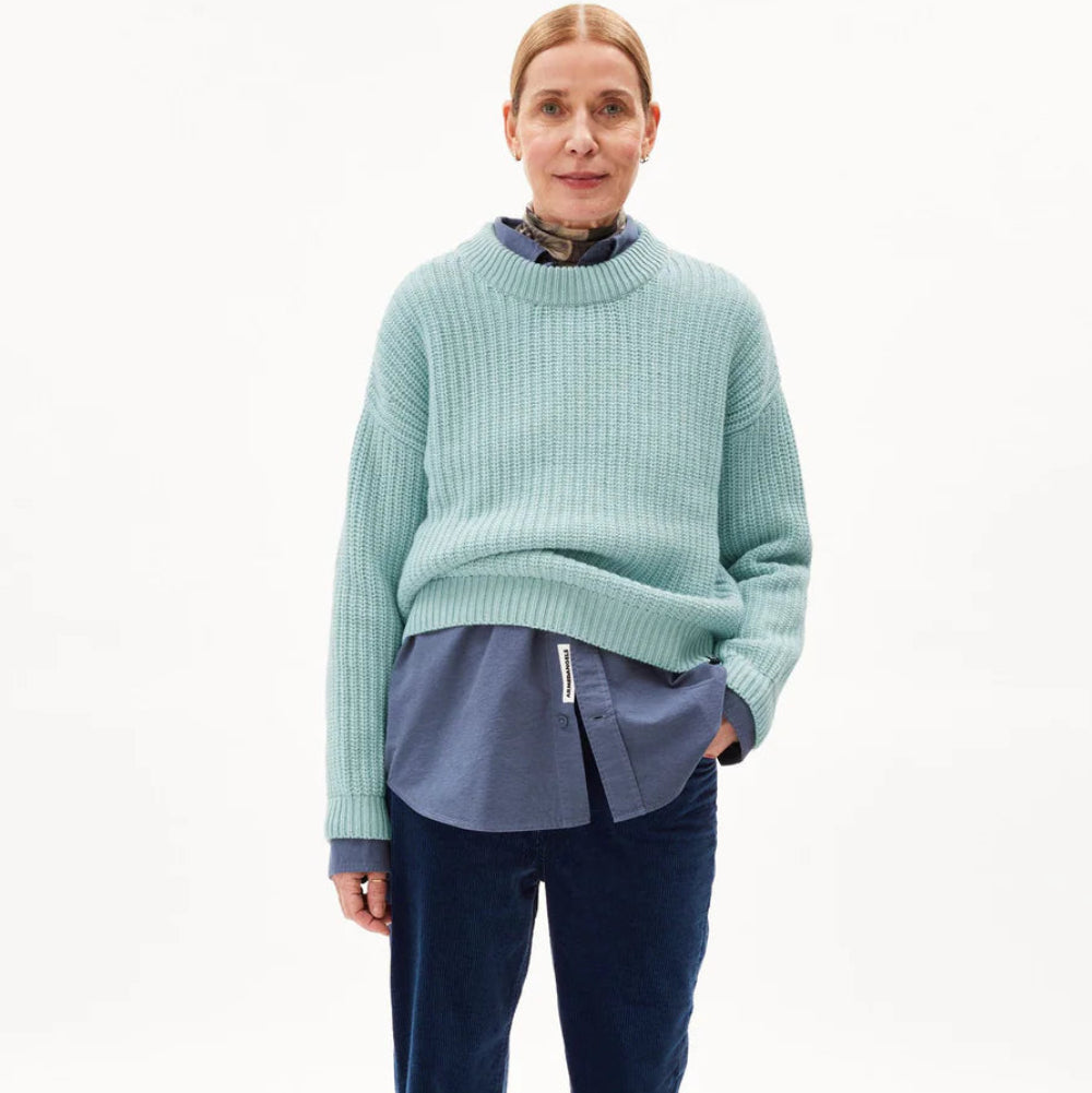 Eine Frau steht vor einem schlichten weißen Hintergrund, trägt einen hellblauen Strickpullover über einem blauen Button-up-Hemd und eine dunkelblaue Hose, die Haare nach hinten gezogen und eine Hand in der Tasche.