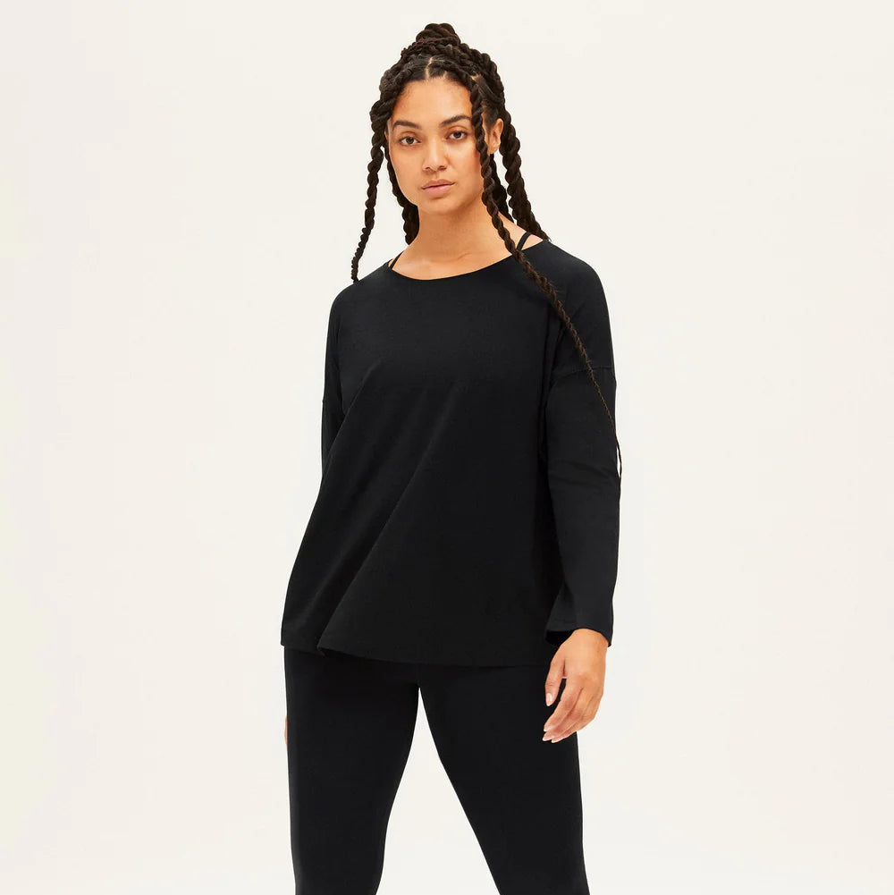 Eine Frau mit langen Zöpfen steht barfuß vor einem schlichten, hellen Hintergrund und trägt ein schwarzes, langärmeliges Oberteil und passende schwarze Leggings.