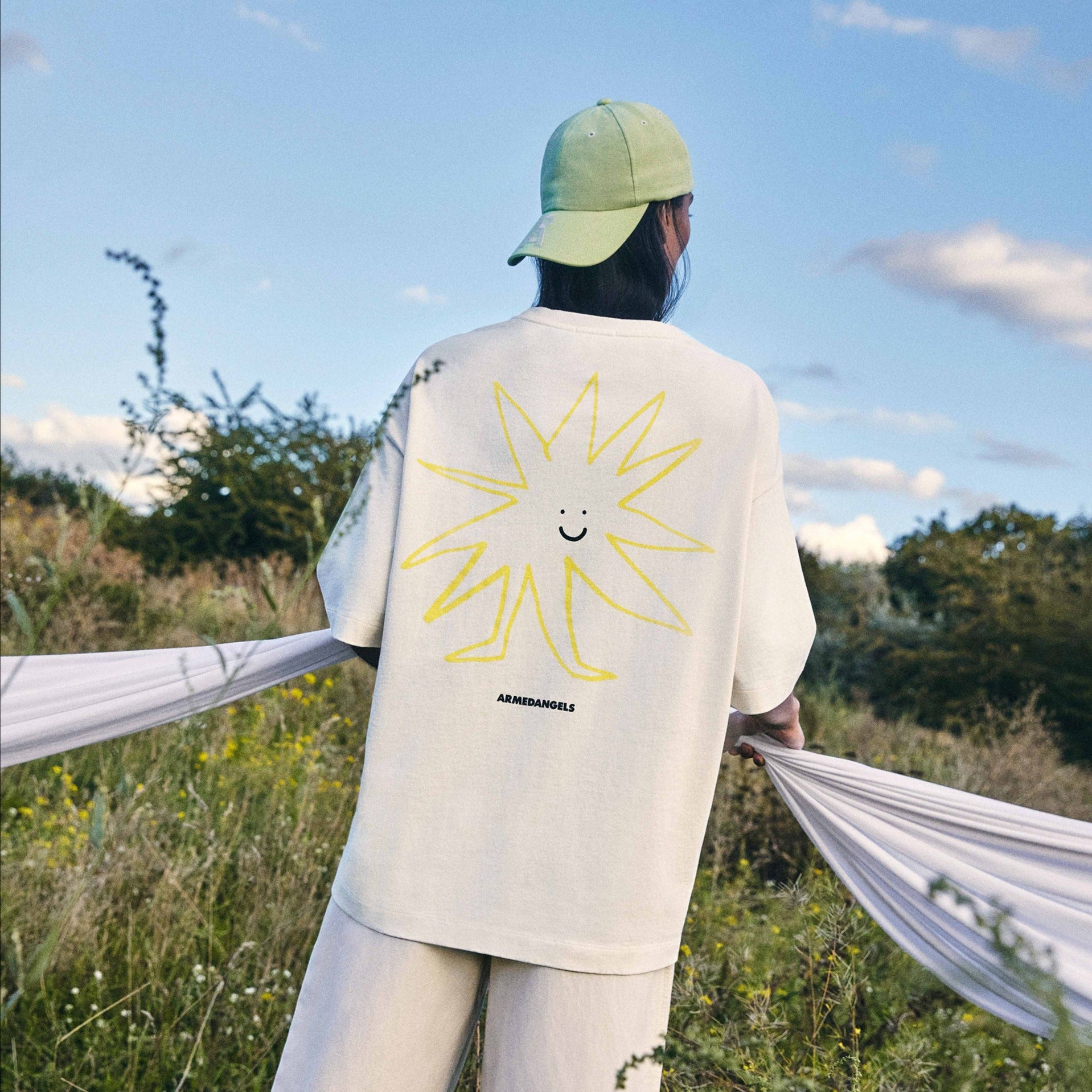 Person mit grünem Basecap trägt ein T-Shirt mit gelbem Lächelndem-Sonnen-Design in einer Wiese bei blauem Himmel.
