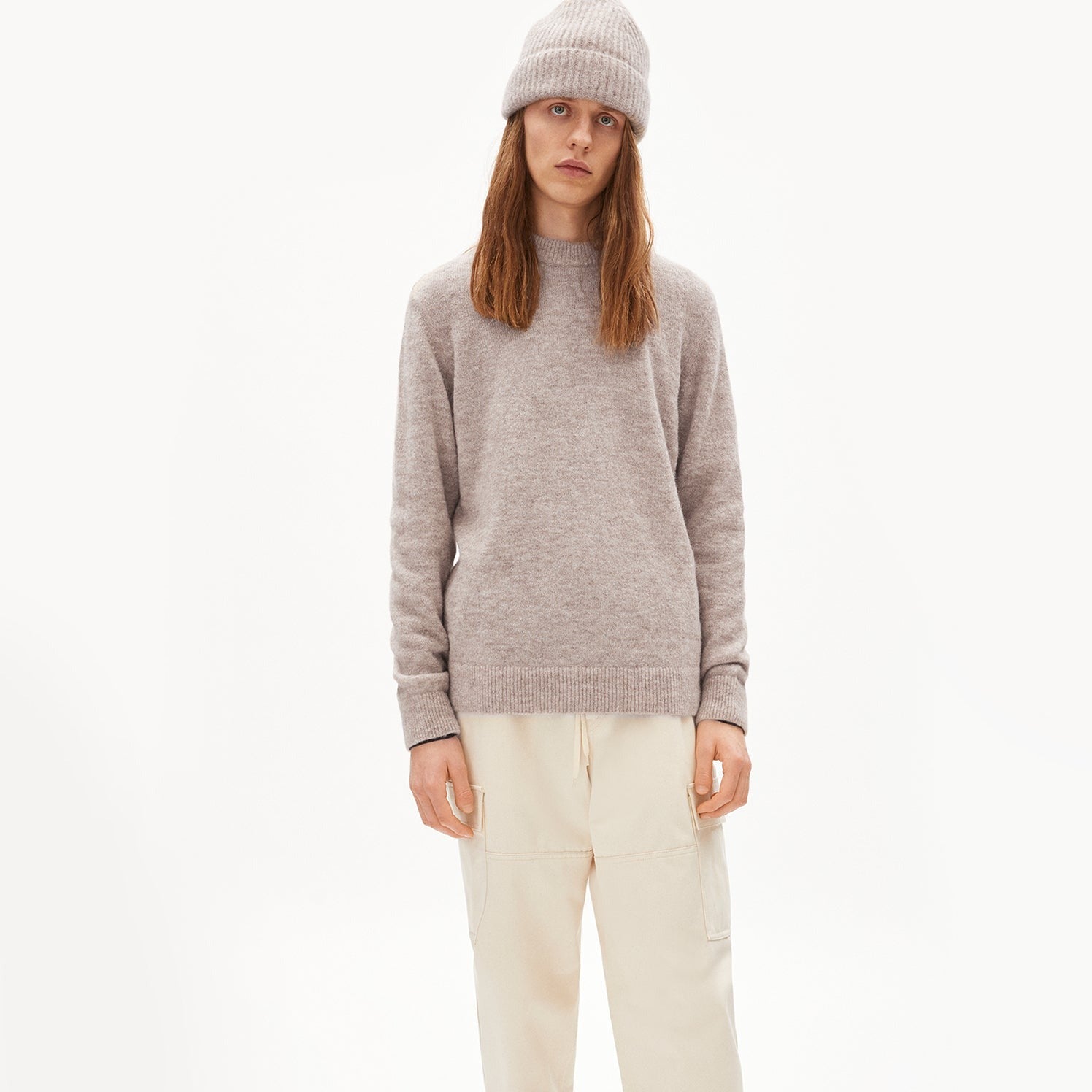 Person mit langen Haaren trägt einen grauen Pullover, eine passende Mütze und beige Hose vor weißem Hintergrund.