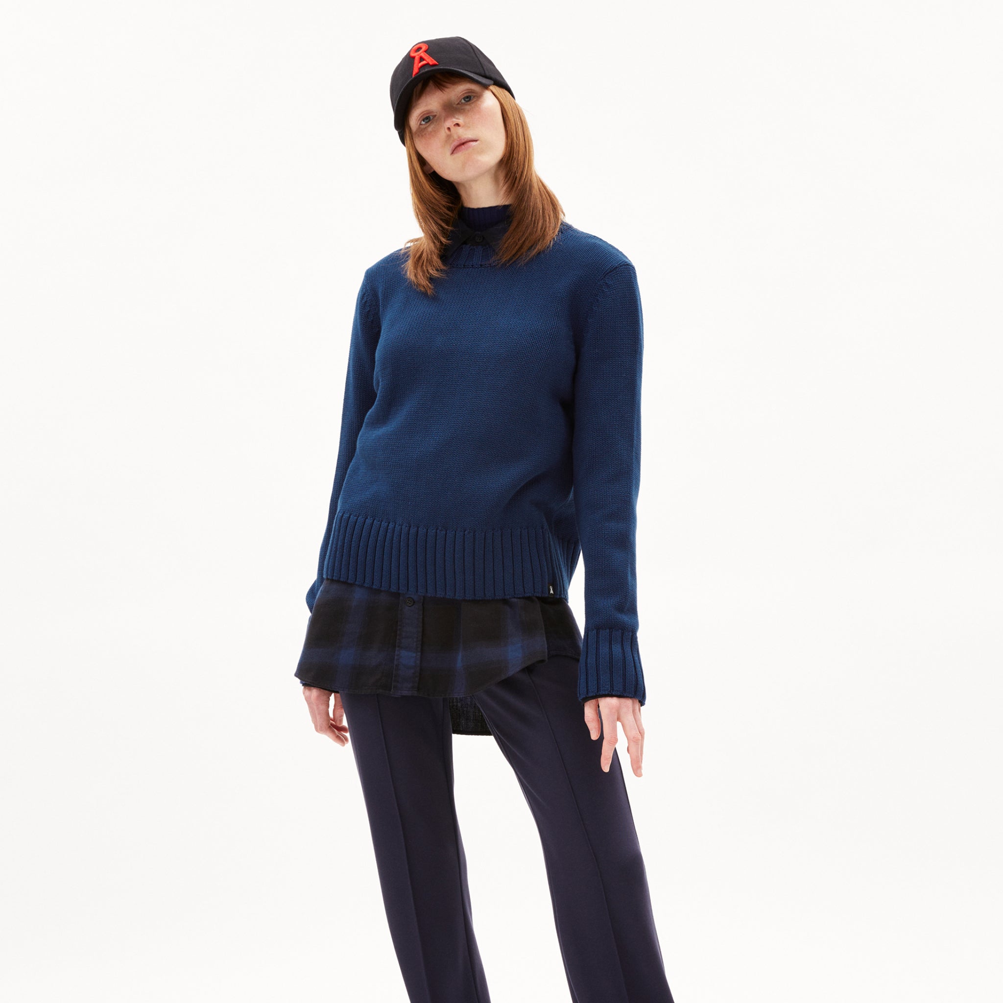 Person mit blauem Pullover, schwarzem Hemd und schwarzer Hose trägt eine schwarze Baseballkappe mit rotem Symbol.