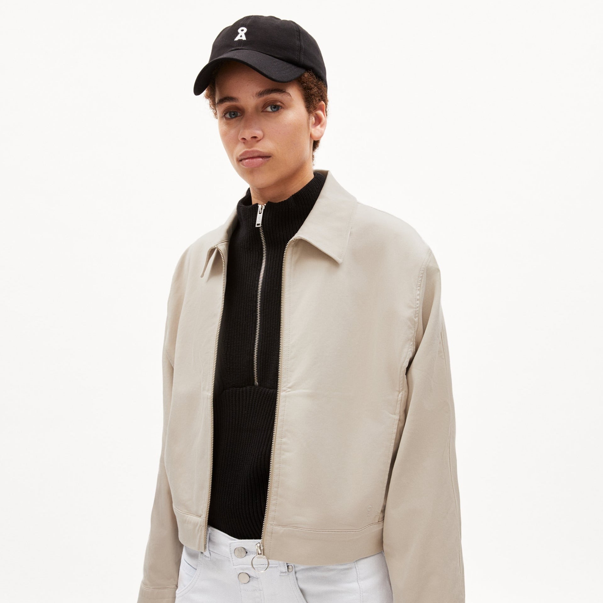 Person in hellgrauer Jacke, schwarzem Pullover mit Reißverschluss, schwarzer Kappe und weißer Hose vor weißem Hintergrund.