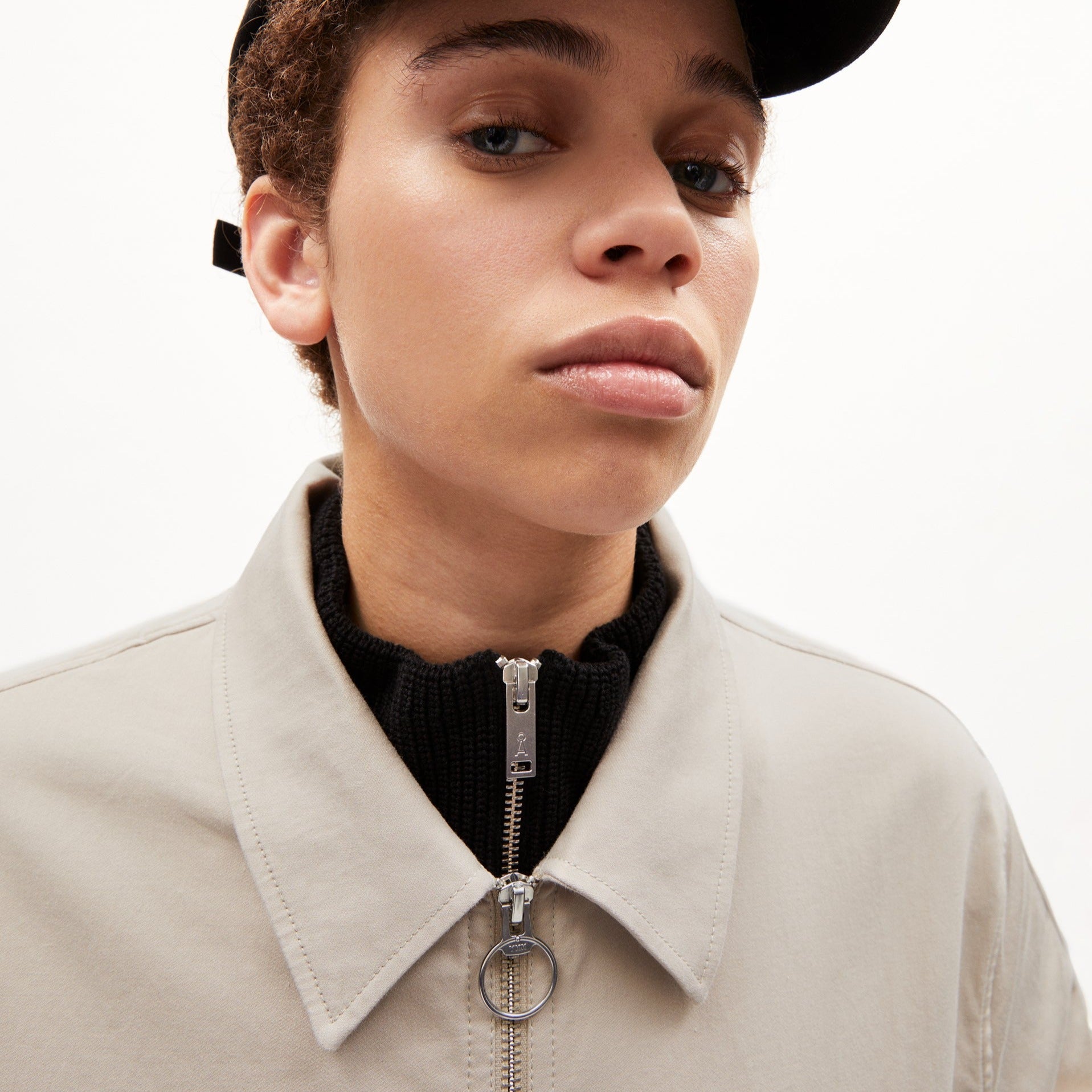 Person mit hellem Mantel und Reißverschluss schaut ernst in die Kamera, trägt eine schwarze Mütze.