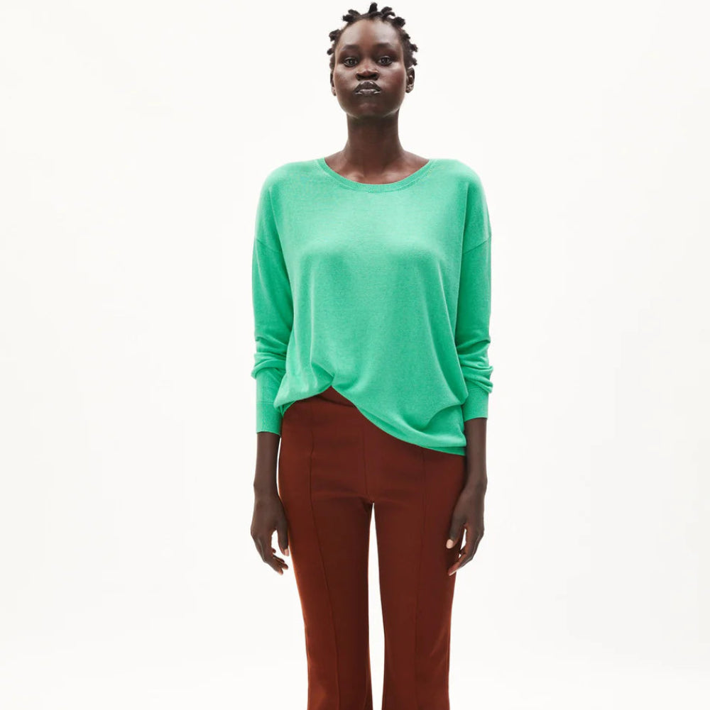 Eine Person steht vor einem schlichten weißen Hintergrund und trägt ein lockeres mintgrünes langärmeliges Oberteil, rostfarbene Schlaghosen und klobige cremefarbene Schuhe.