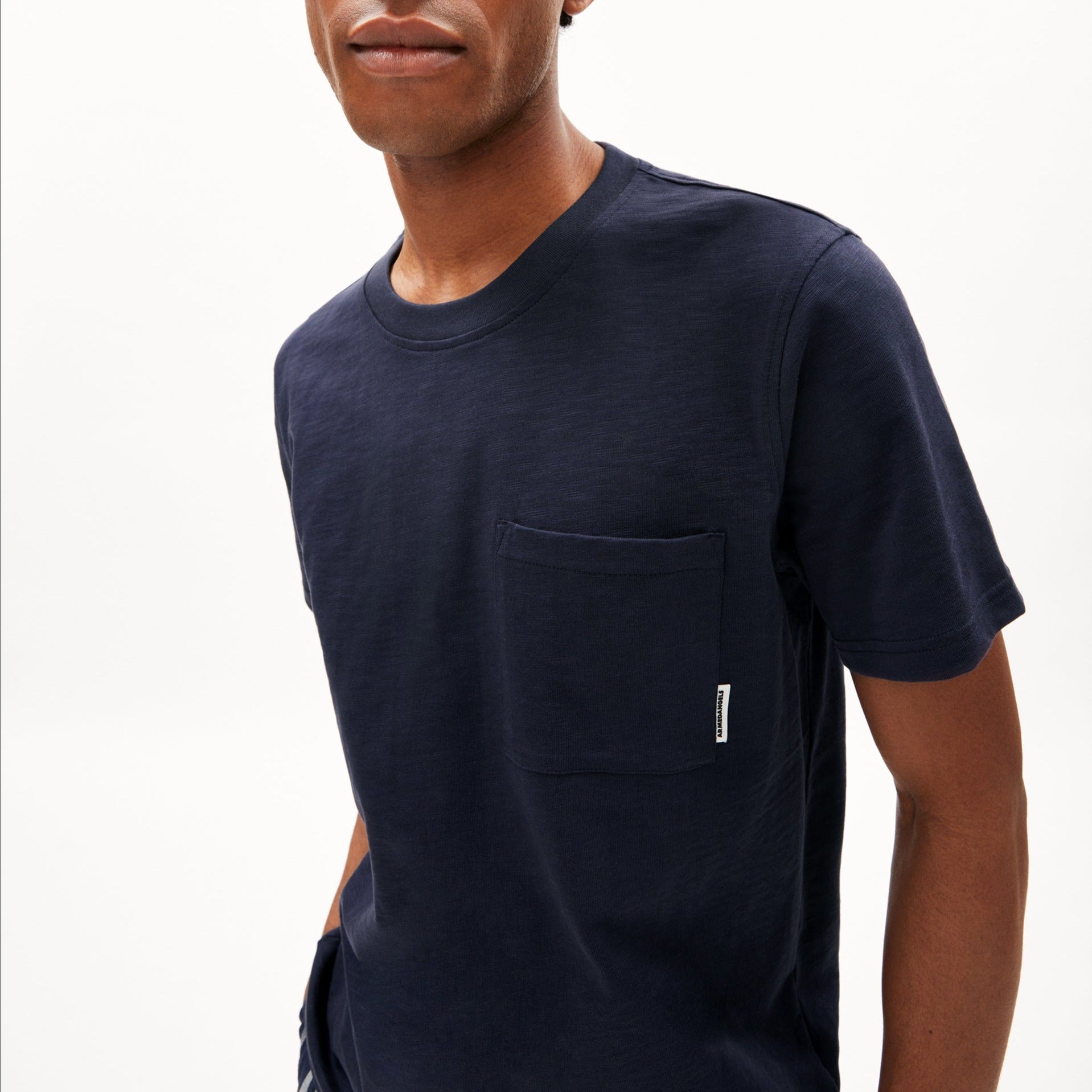 Person in dunkelblauem T-Shirt mit Brusttasche vor weißem Hintergrund.