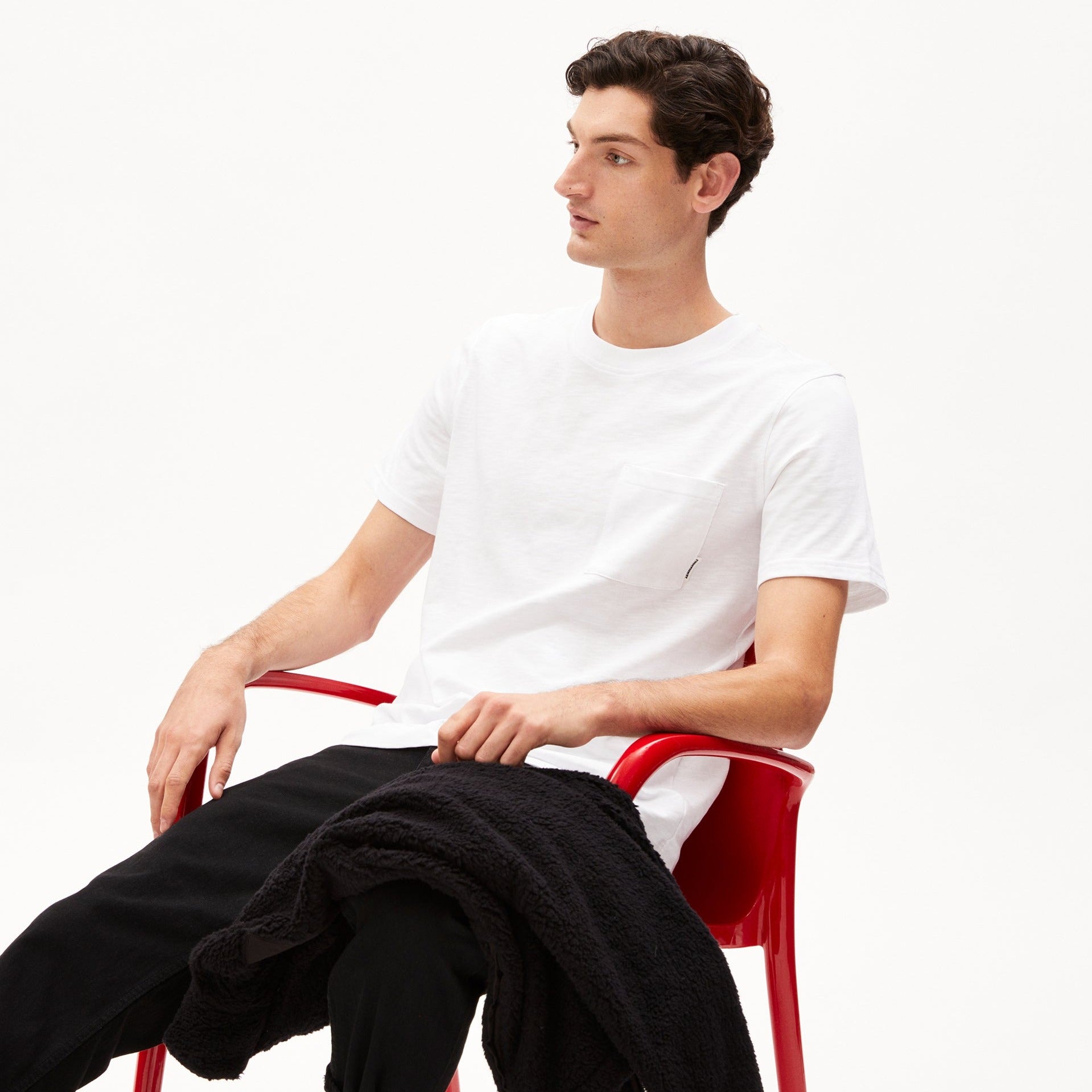 Junger Mann sitzt entspannt in einem roten Stuhl und trägt ein weißes T-Shirt und eine schwarze Hose.
