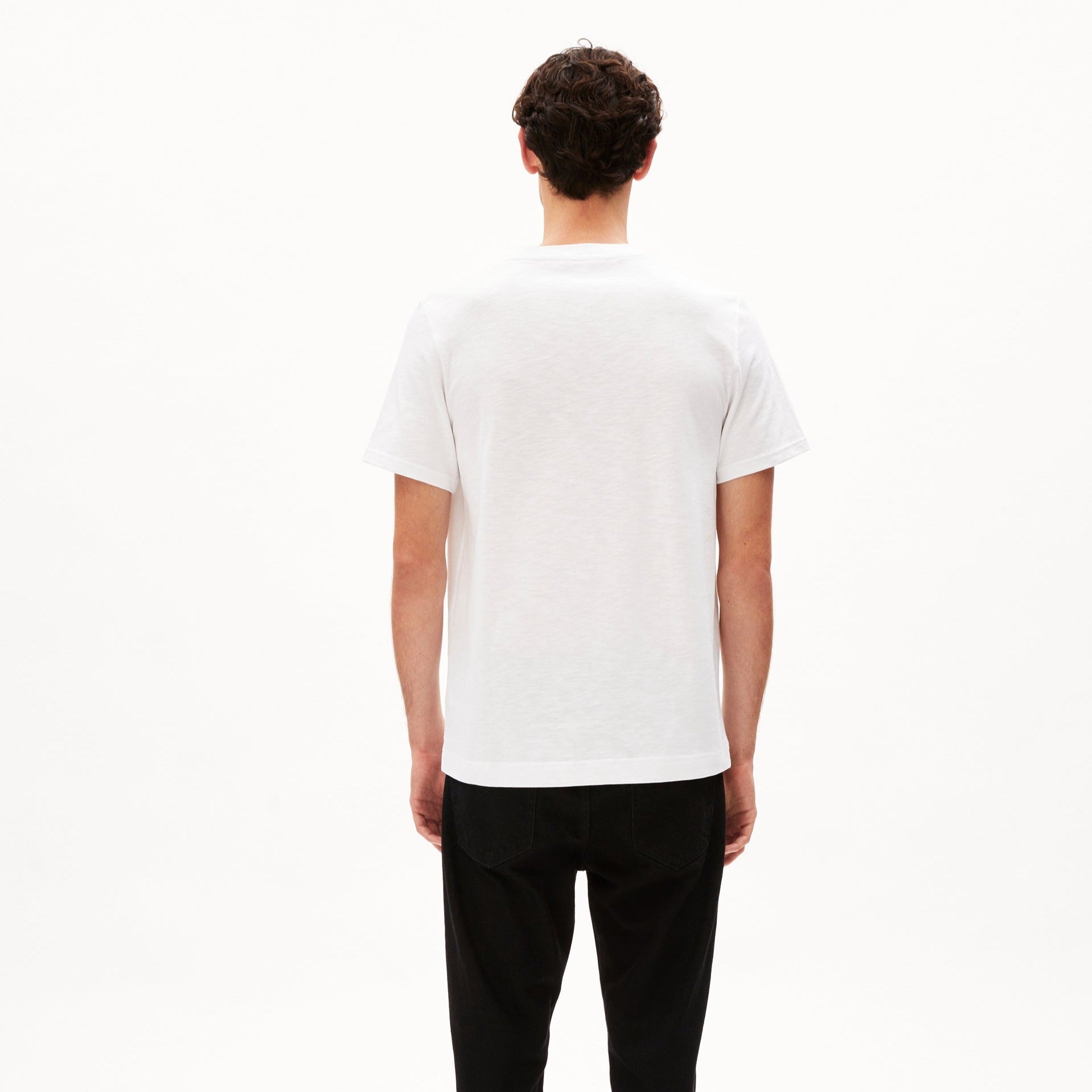 Mann in weißem T-Shirt und dunkler Hose steht mit dem Rücken zur Kamera vor einem weißen Hintergrund.