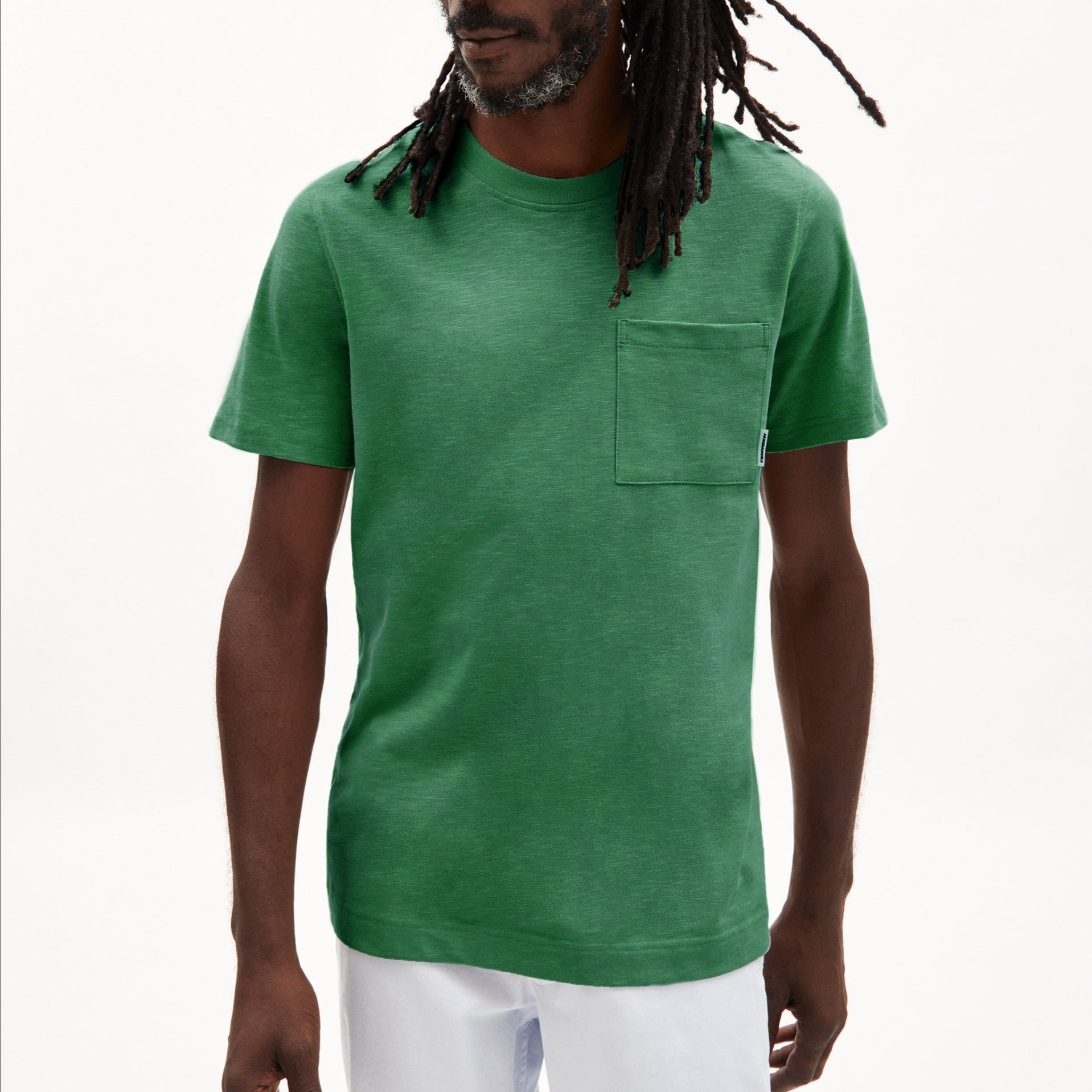 Mann mit lockigen Haaren trägt ein grünes T-Shirt mit Brusttasche und weiße Hose vor einem hellen Hintergrund.