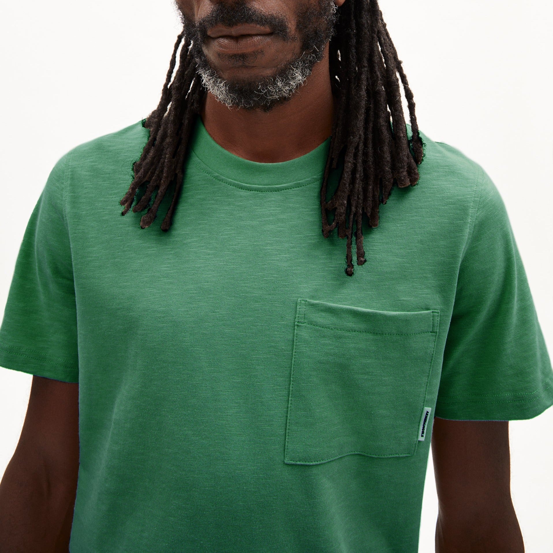 Person mit langen Dreadlocks trägt ein grünes T-Shirt mit Brusttasche vor einem neutralen Hintergrund.
