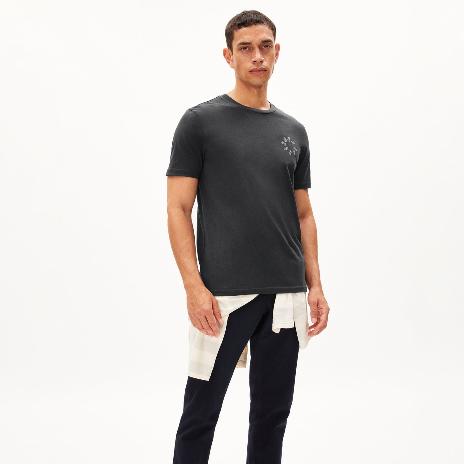 Mann in schwarzem T-Shirt mit aufgedrucktem Muster, weißes Hemd um die Taille gebunden, schwarze Hose, neutraler Hintergrund.