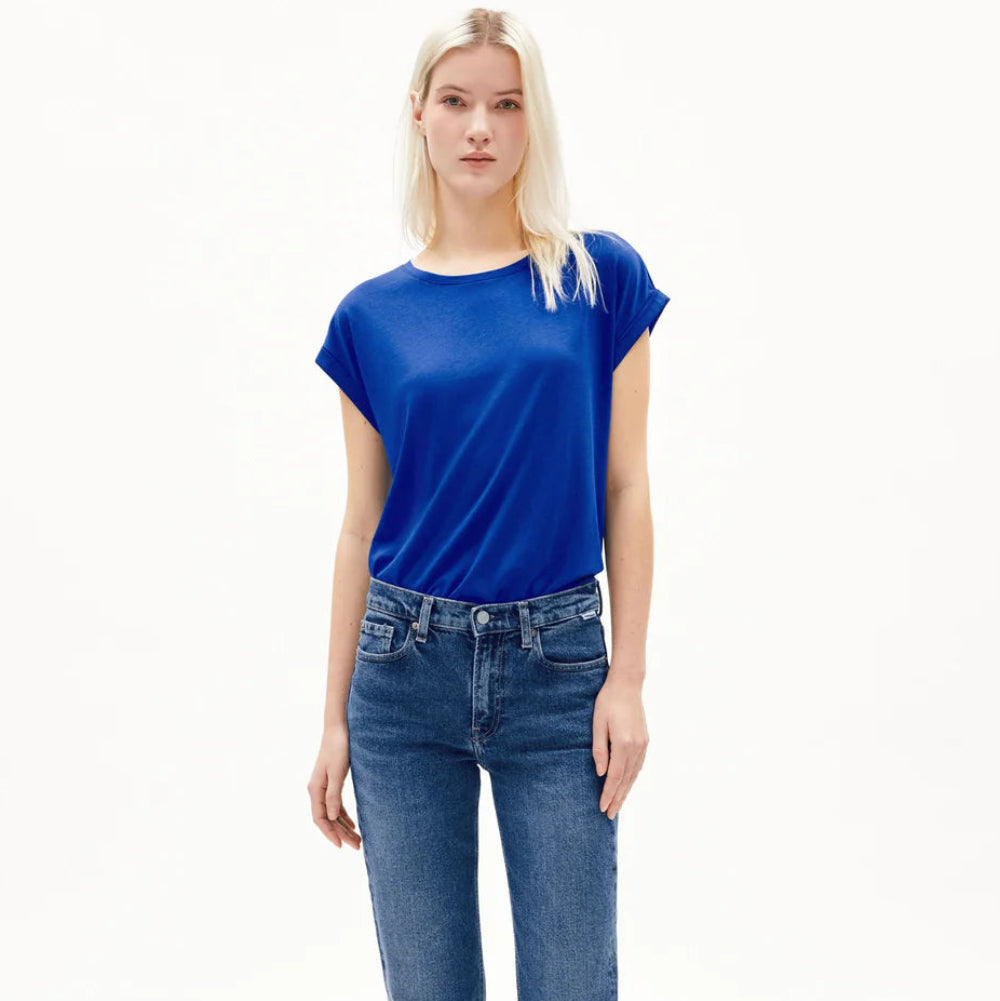 Eine Frau mit langen blonden Haaren trägt ein hellblaues T-Shirt, eine Jeans mit geradem Bein und weiße Schuhe und steht vor einem schlichten weißen Hintergrund.