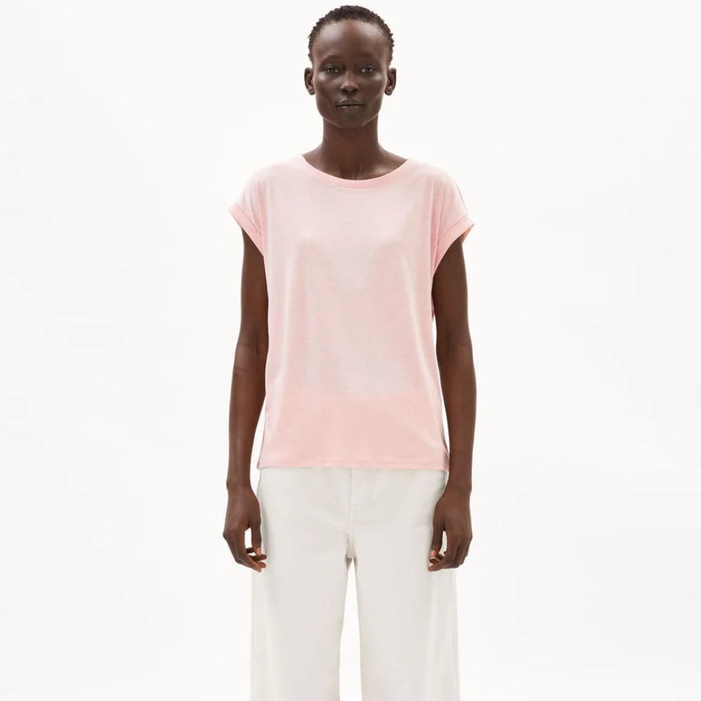 Eine Person, die ein hellrosa kurzärmeliges Oberteil und eine locker sitzende weiße Hose mit weitem Bein trägt, steht vor einem schlichten weißen Hintergrund.