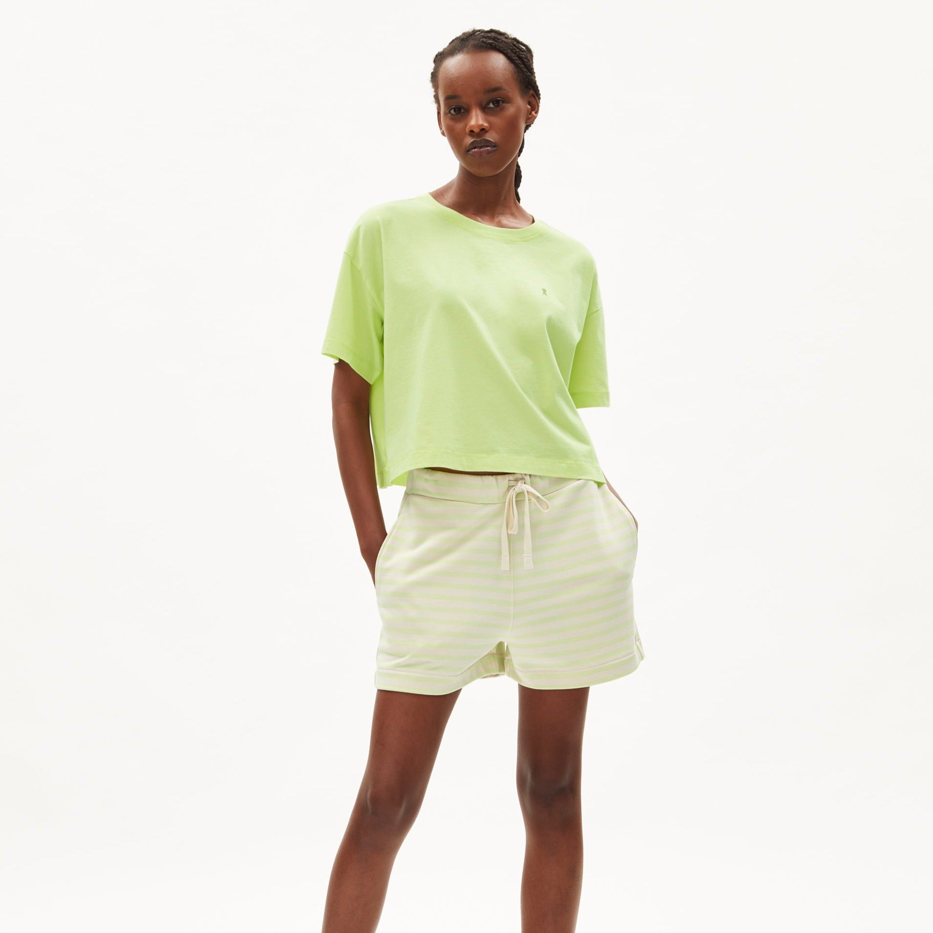 Frau trägt ein hellgrünes T-Shirt und gestreifte Shorts, steht vor weißem Hintergrund.