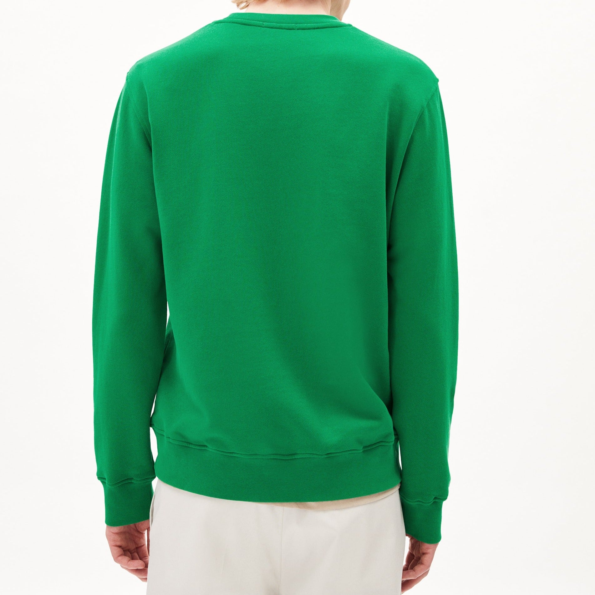 Person mit einem grünen Pullover von hinten, trägt beige Hosen vor einem weißen Hintergrund.