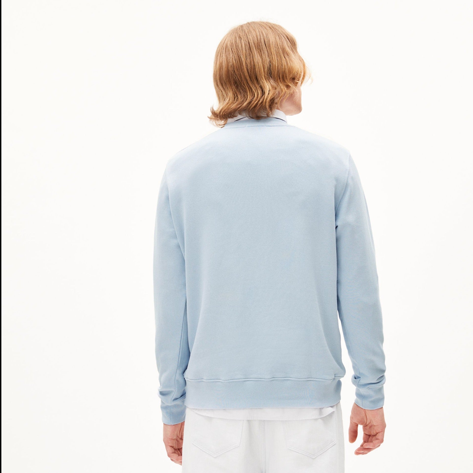 Rückansicht einer Person mit schulterlangem blonden Haar in einem hellblauen Pullover und weißer Hose vor weißem Hintergrund.