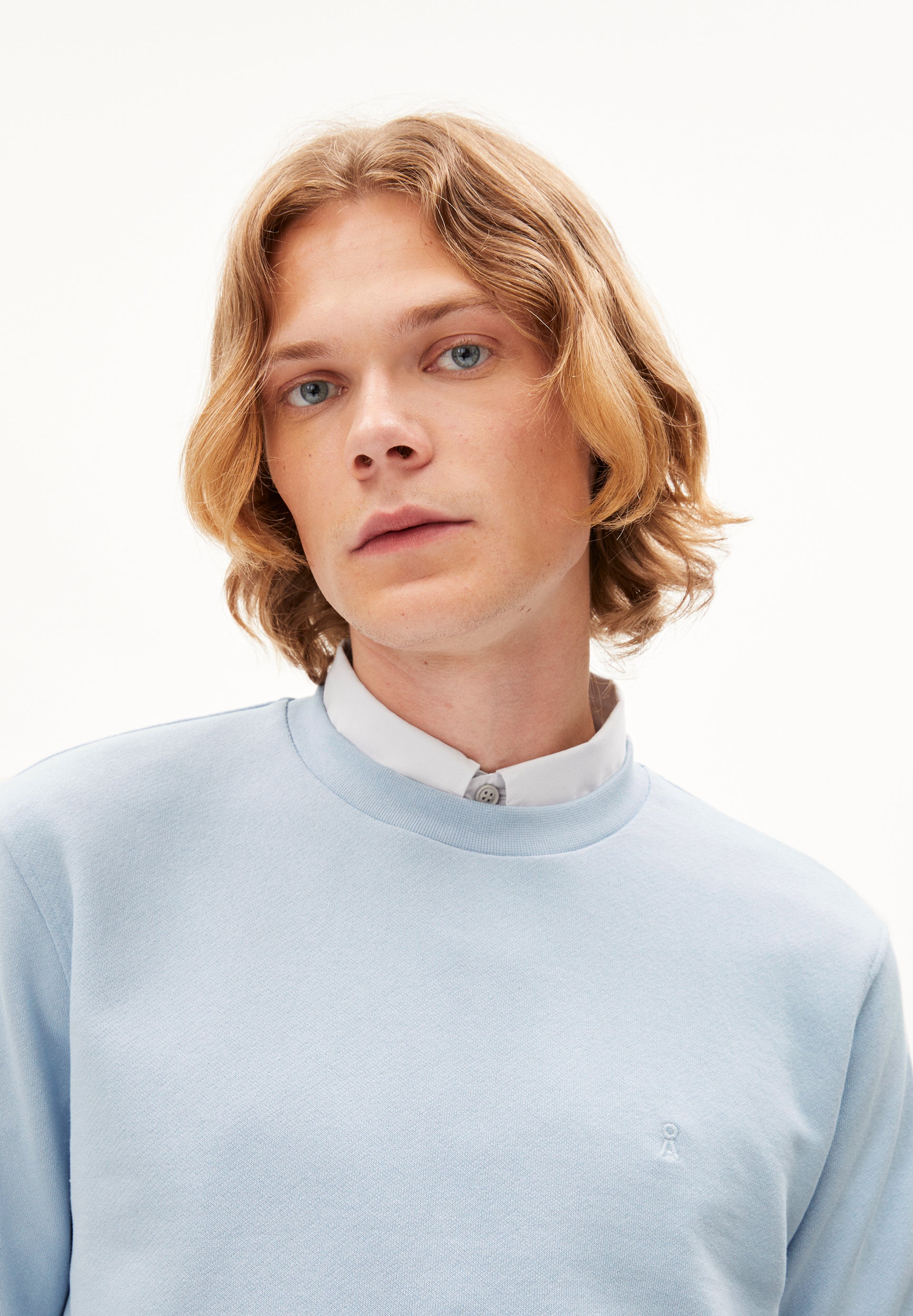 Blonde Person in light blue sweater and white collared shirt, looking at the camera against a plain white background.