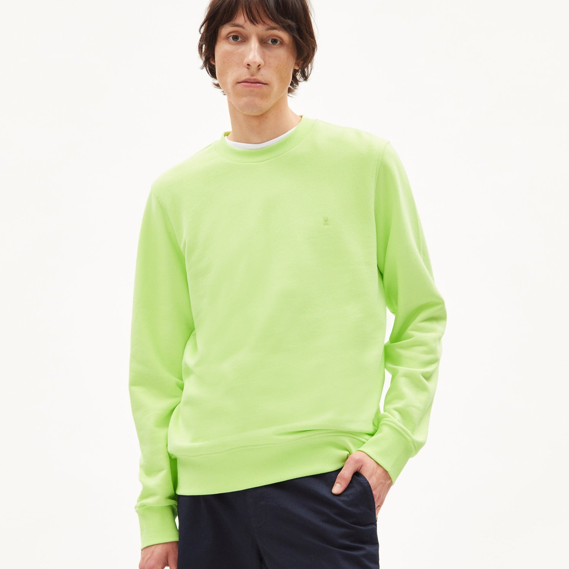 Person in einem hellgrünen Pullover und dunklen Hosen vor einem weißen Hintergrund.