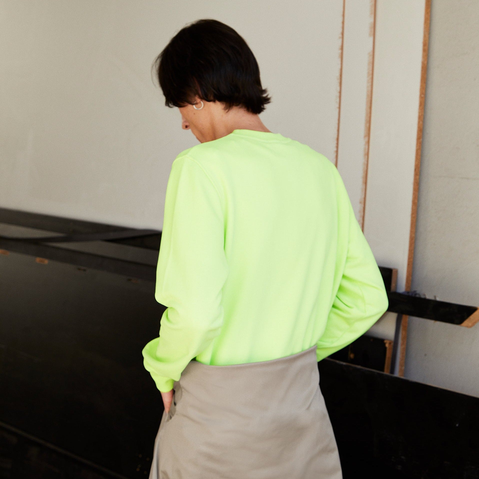 Person mit neonfarbenem Pullover und grauer Hose steht in einem Innenraum mit dem Rücken zur Kamera.