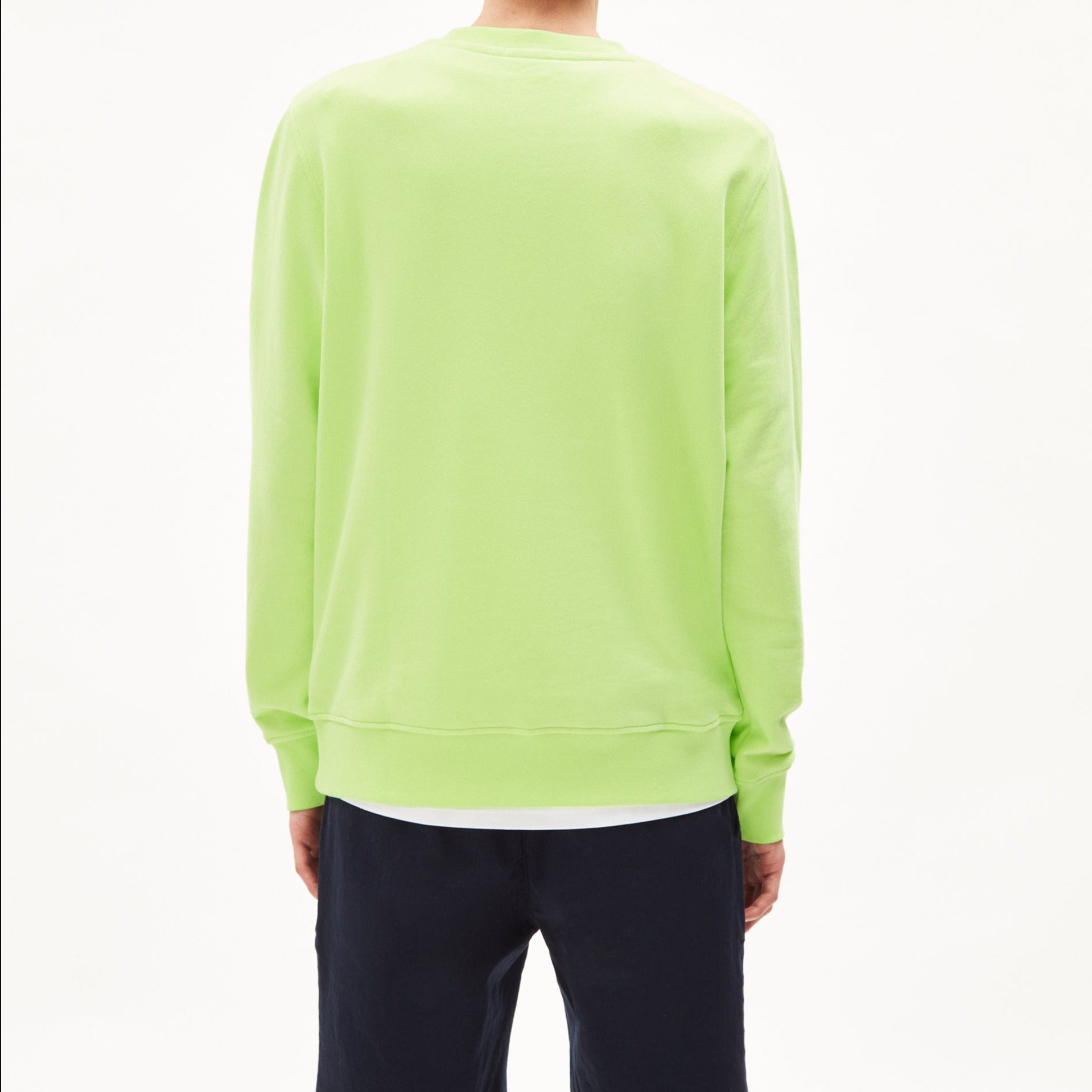 Eine Person steht mit dem Rücken zur Kamera in einem hellgrünen Pullover und schwarzen Hosen vor einem weißen Hintergrund.