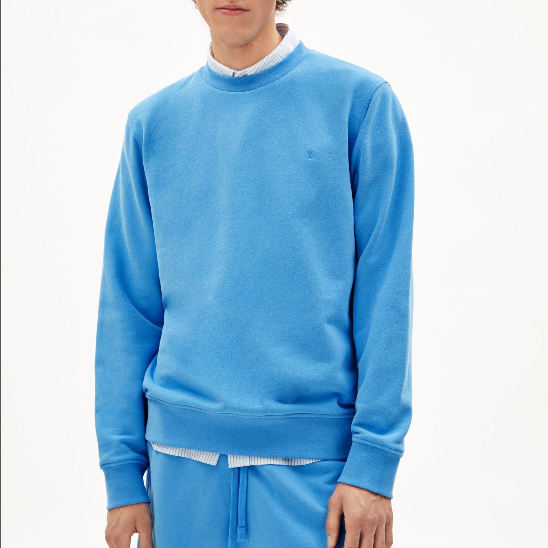 Ein Mann trägt einen blauen Pullover und passende Jogginghose vor weißem Hintergrund.