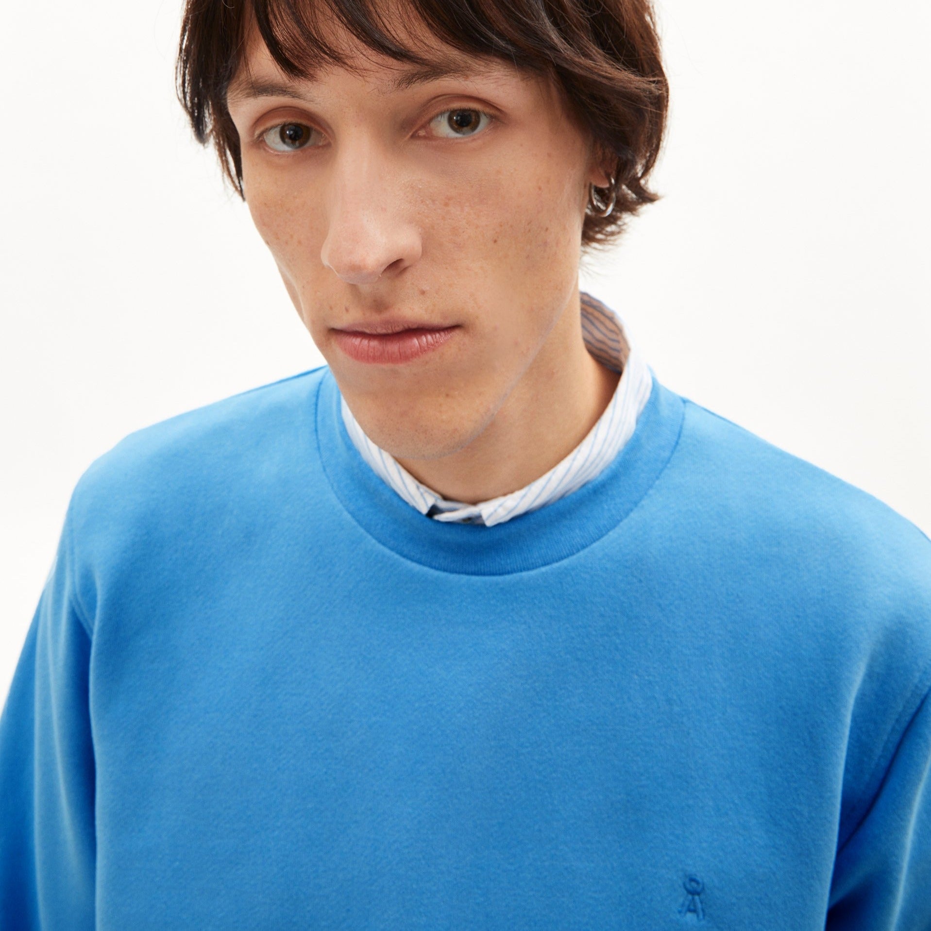 Ein Mann trägt einen blauen Pullover über einem gestreiften Hemd und schaut direkt in die Kamera.