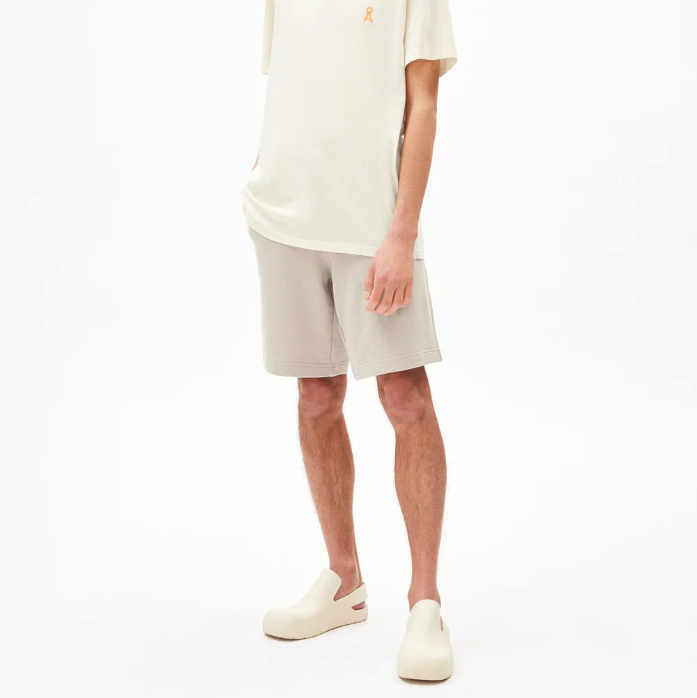 Ein junger Mann mit blassgelber Mütze, weißem T-Shirt, hellbeigen Shorts und cremefarbenen Slippern steht vor einem schlichten weißen Hintergrund und hat die Hände in den Taschen.